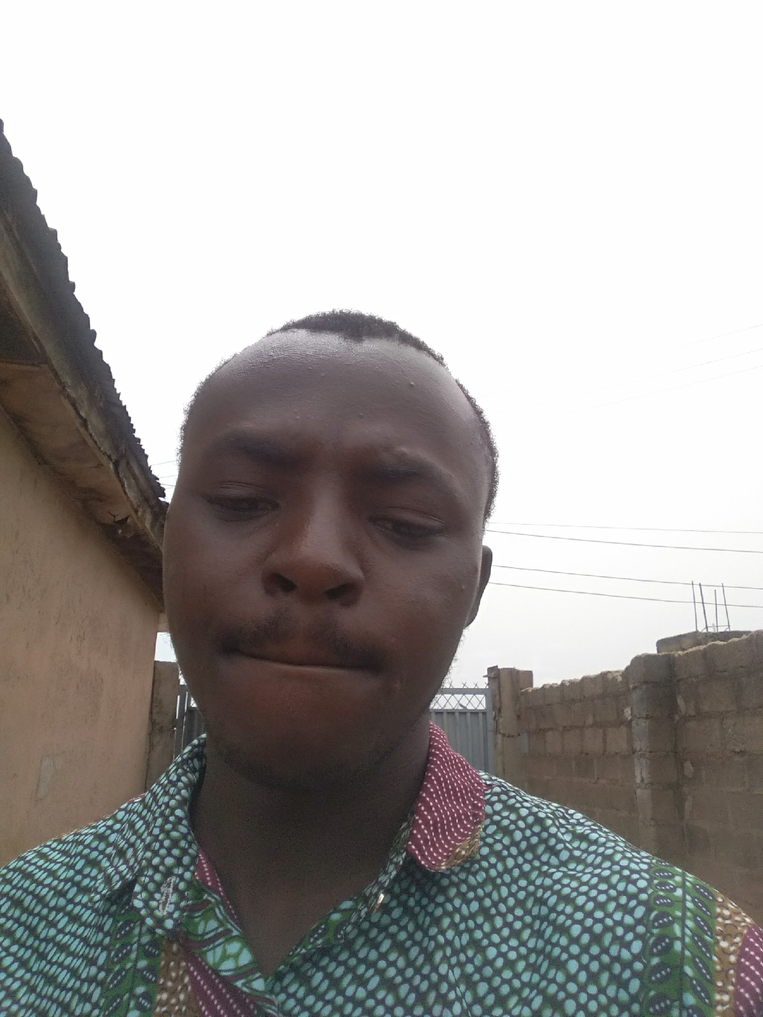 The image shows a medium shot of a man with dark skin, a mustache, and short hair. He is wearing a patterned shirt with a green and white polka dot design, burgundy accents, and hints of brown and green. The background is varied, showing a beige building to the left with a visible roof line, a gray overcast sky, and a partially constructed cinder block wall on the right. Power lines are visible in the upper right corner. The location is the city of Unknown, Unknown.