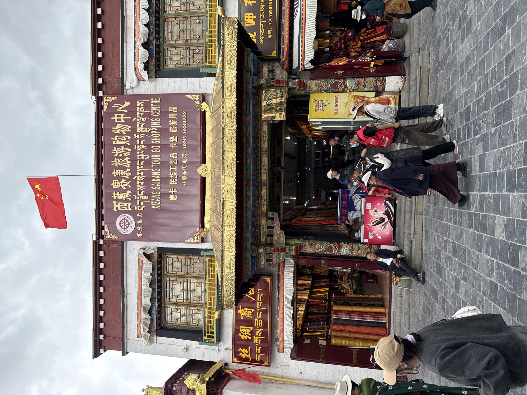 Here's a detailed description of the image:
**Overview:**
The image captures a street scene in what appears to be a commercial area, possibly a shopping or tourist district, in the city of Chengguan District, China. The buildings feature traditional architectural elements.
**Environment:**
The street is paved with stone or brick, and the sky is overcast. There are shop fronts with goods displayed, indicating a bustling commercial activity. The general atmosphere seems lively due to the people present.
**Buildings and Architecture:**
The buildings are multi-story structures. The facades have traditional designs with golden accents, decorative eaves, and windows with traditional frames. One building has a prominent sign in both Chinese and Tibetan. There is also a sign visible in Chinese characters advertising silk products. The Chinese flag is prominently displayed on one of the buildings.
**People:**
Several people are present in the scene, mostly pedestrians walking or standing near the shops. Some are wearing hats, and some have facemasks. One individual is sitting on a chair.
**Objects:**
Various items are displayed in the shop fronts, including textiles, carpets, and what appear to be souvenirs. There are signs indicating the types of products available. A refrigerator with a drink product is also visible.
**Actions and Interactions:**
People are walking past shops, likely browsing or shopping. There are signs that goods are being traded here. The interactions are typical of a busy market or shopping street.