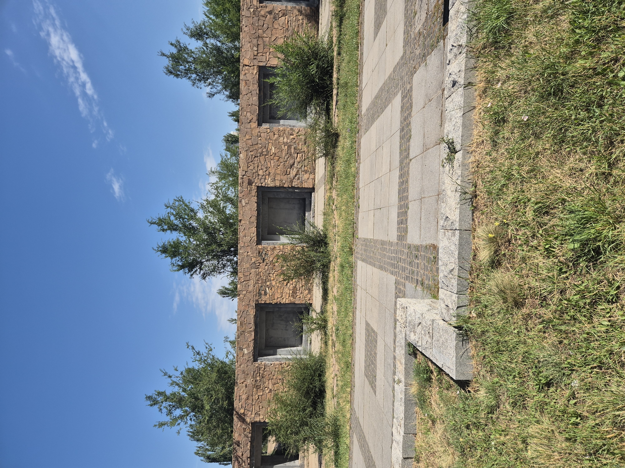 A wide shot captures a stone wall with multiple openings in Astana, Kazakhstan, on a bright, sunny day. The wall, constructed of rough-hewn, light brown stones, features several recessed rectangular openings that appear to be empty. Small, dense green bushes grow in front of some of these openings, and the ground in front of the wall is covered in dry, yellowing grass.
In the foreground, a paved pathway is visible. It consists of large, light gray square tiles arranged in a grid pattern, interspersed with smaller, darker gray cobblestones in a decorative border. The pathway is bordered by a low stone structure made of larger, gray rectangular blocks. More dry grass and sparse green vegetation grow around this structure.
Behind the stone wall, a line of trees with dense green foliage stretches across the frame, silhouetted against a clear, bright blue sky with a few wispy white clouds. The overall scene suggests an outdoor public space, possibly a park or memorial, characterized by its man-made structures and natural elements.