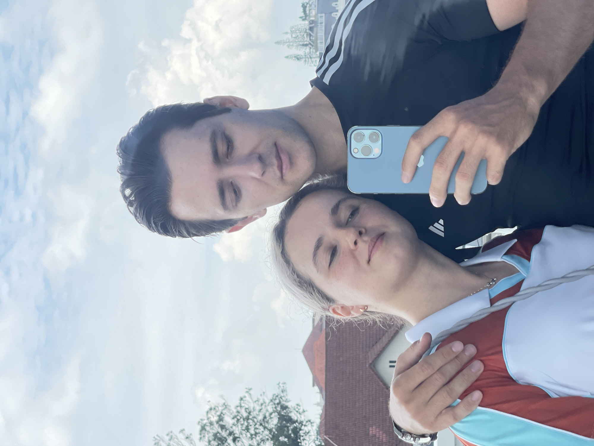 A man and a woman are taking a selfie. The man is wearing a black Adidas shirt with white stripes on the sleeves. He is holding a light blue iPhone with three camera lenses. The woman is wearing a white, red, and teal sports top. The background shows a cloudy sky and part of a building with a red roof. Based on the style of the building, this is most likely not in Lâm Viên - Đà Lạt, Vietnam, which is renowned for its distinct French colonial architecture. The location is probably in Europe.
