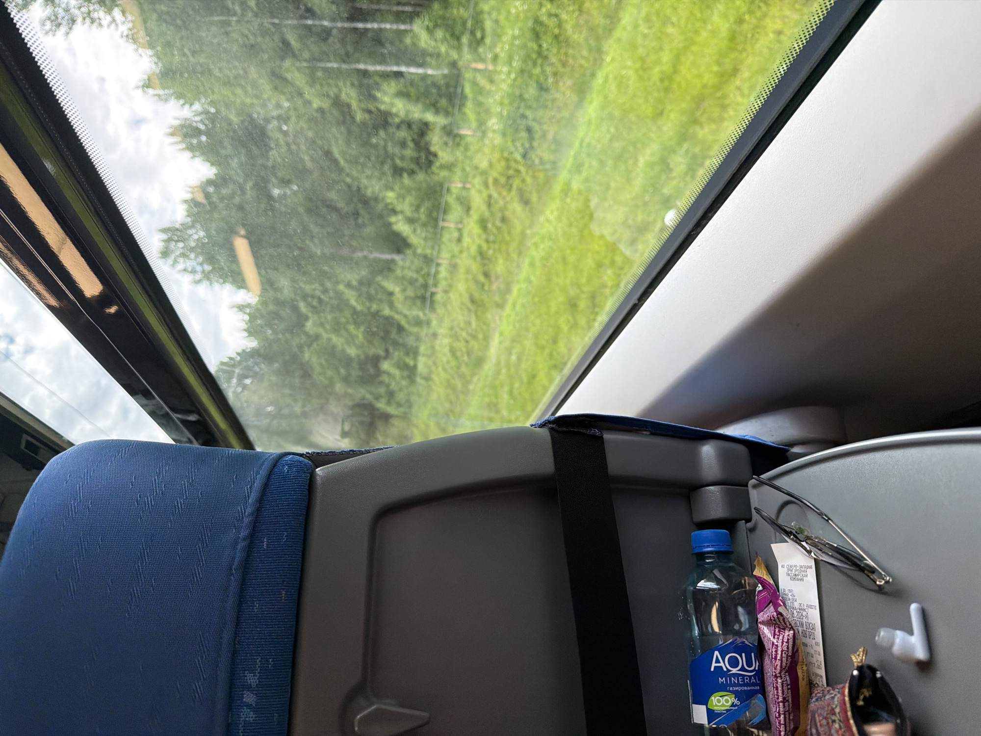 This media file captures a view from inside a train, looking out a large window. The main subjects are the train's interior elements, including a blue fabric-covered seat on the left and a gray plastic seatback with various items attached to it on the right. The window offers a scenic view of a lush green landscape with trees and grass, suggesting an outdoor setting. The time of day appears to be daytime, with bright lighting filtering through the window, and the sky, visible at the top left, appears partly cloudy.
On the right side of the seatback, several objects are visible. These include a bottle of "AQUA MINERAL" water, a packet of what appears to be a snack with text that includes "Raisins And Caramelx," a pair of eyeglasses, and a receipt. The receipt displays some text in Cyrillic, with a snippet readable as "АО СЕВЕРО-ЗАПАДНАЯ ПРИГОРОДНАЯ ПАССАЖИРСКАЯ," indicating it might be from a regional passenger company in Russia. There's also a small white hook attached to the seatback.
The overall impression is a snapshot of a journey on a train, likely through a rural or natural environment. The focus is on the immediate surroundings within the train car and the passing scenery outside. No people are visible in the frame.