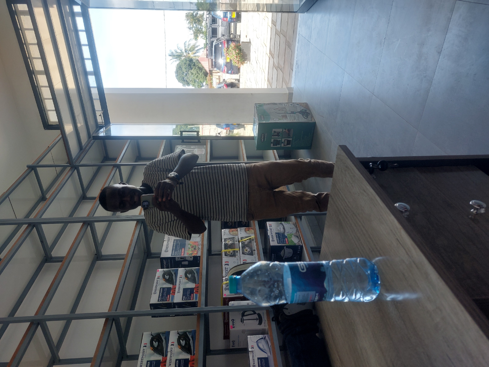 A man is taking a selfie in an appliance store in Dar es-Salaam, Tanzania. He is standing in front of shelves stocked with boxed appliances, including blenders, irons, and kettles. In the foreground, a bottle of water sits on a counter. Outside the store, vehicles are parked on the street.