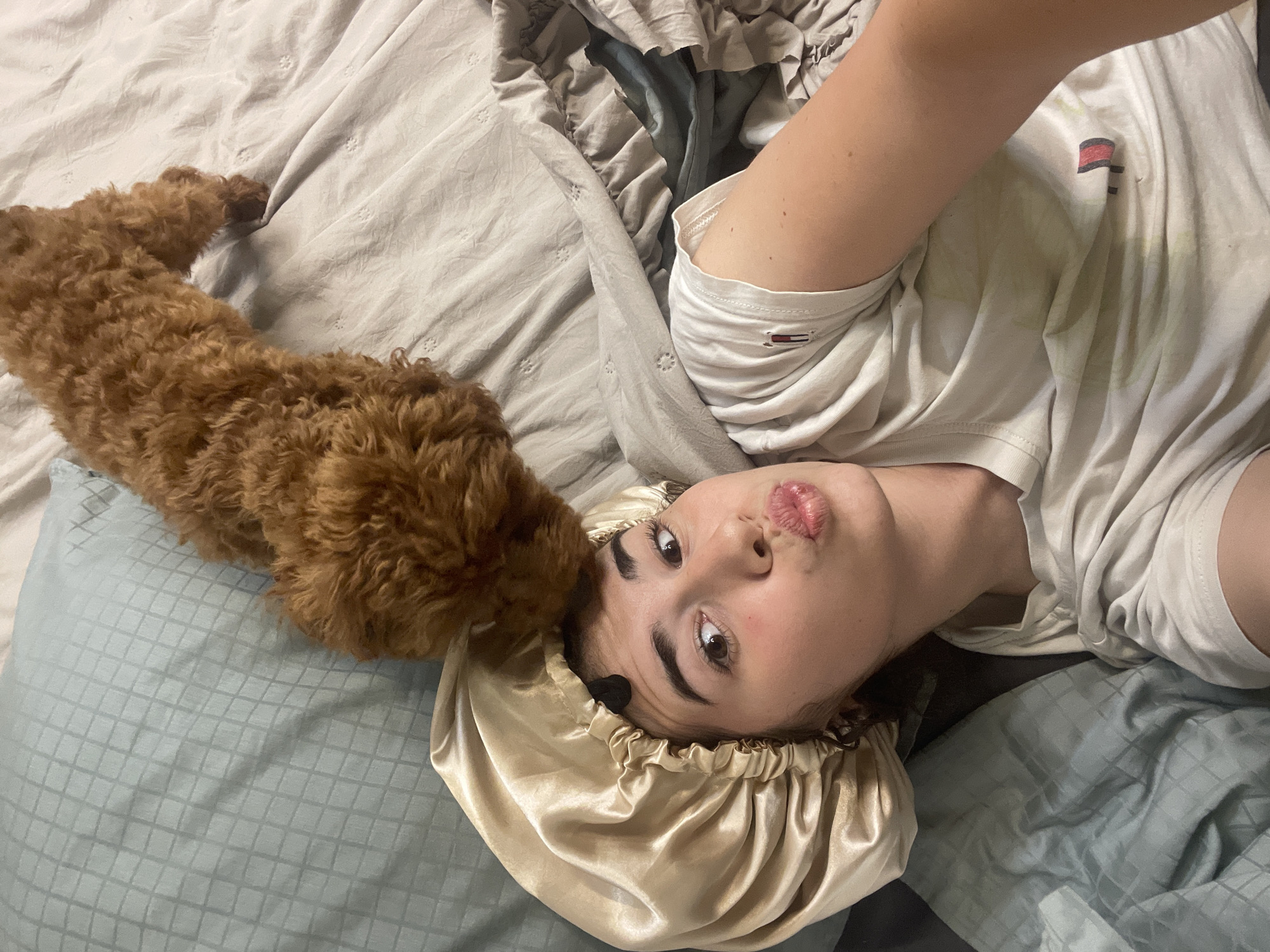 The image depicts a casual indoor scene, likely in a bedroom, featuring a young person and a dog on a bed. The person, appearing to be female, is lying on their back with their head turned towards the camera, suggesting a selfie. They are wearing a light-colored t-shirt with a small Tommy Hilfiger flag logo visible on the left sleeve and a golden-colored satin bonnet on their head. Their lips are pursed in a playful or "pouty" expression.
Next to the person's head is a small, fluffy, reddish-brown dog, resembling a poodle or doodle breed. The dog's head is close to the person's temple, appearing to sniff or gently interact with them. The bed is covered with light beige or off-white textured sheets and light blue-grey pillows. The lighting is soft, indicating an indoor setting. The scene conveys a relaxed and affectionate moment between the person and their pet. The location context is Sofia, Bulgaria.