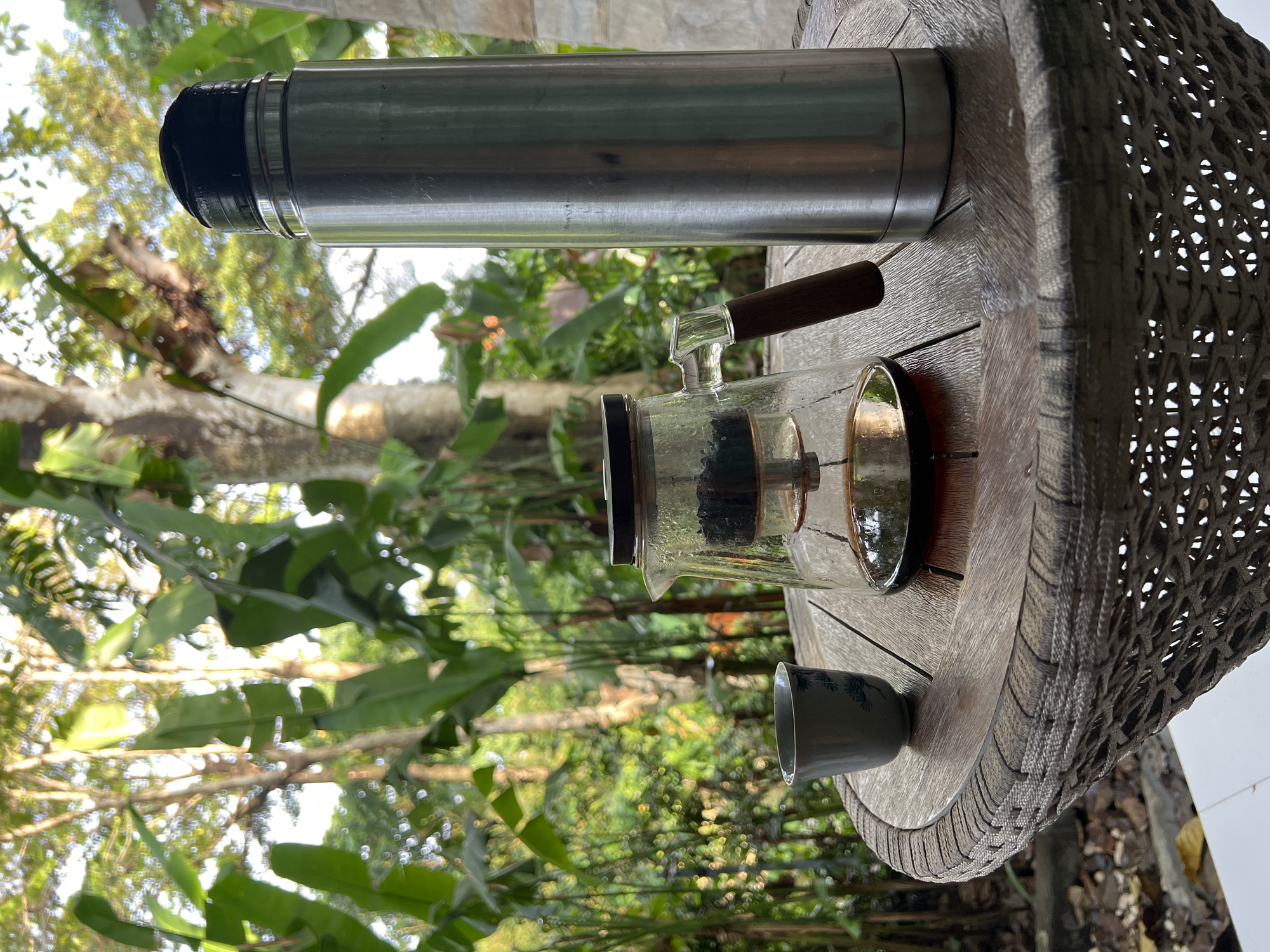 On a round, wooden-surface table with a woven edge, a stainless steel thermos lies horizontally at the top, a glass coffee maker with a dark brown wooden handle is centered, and a small blue cup sits near the bottom. The table appears to be outdoors, set against a backdrop of lush green foliage and trees. The photograph was taken in Kerobokan, Indonesia. There are no people visible in the image.
