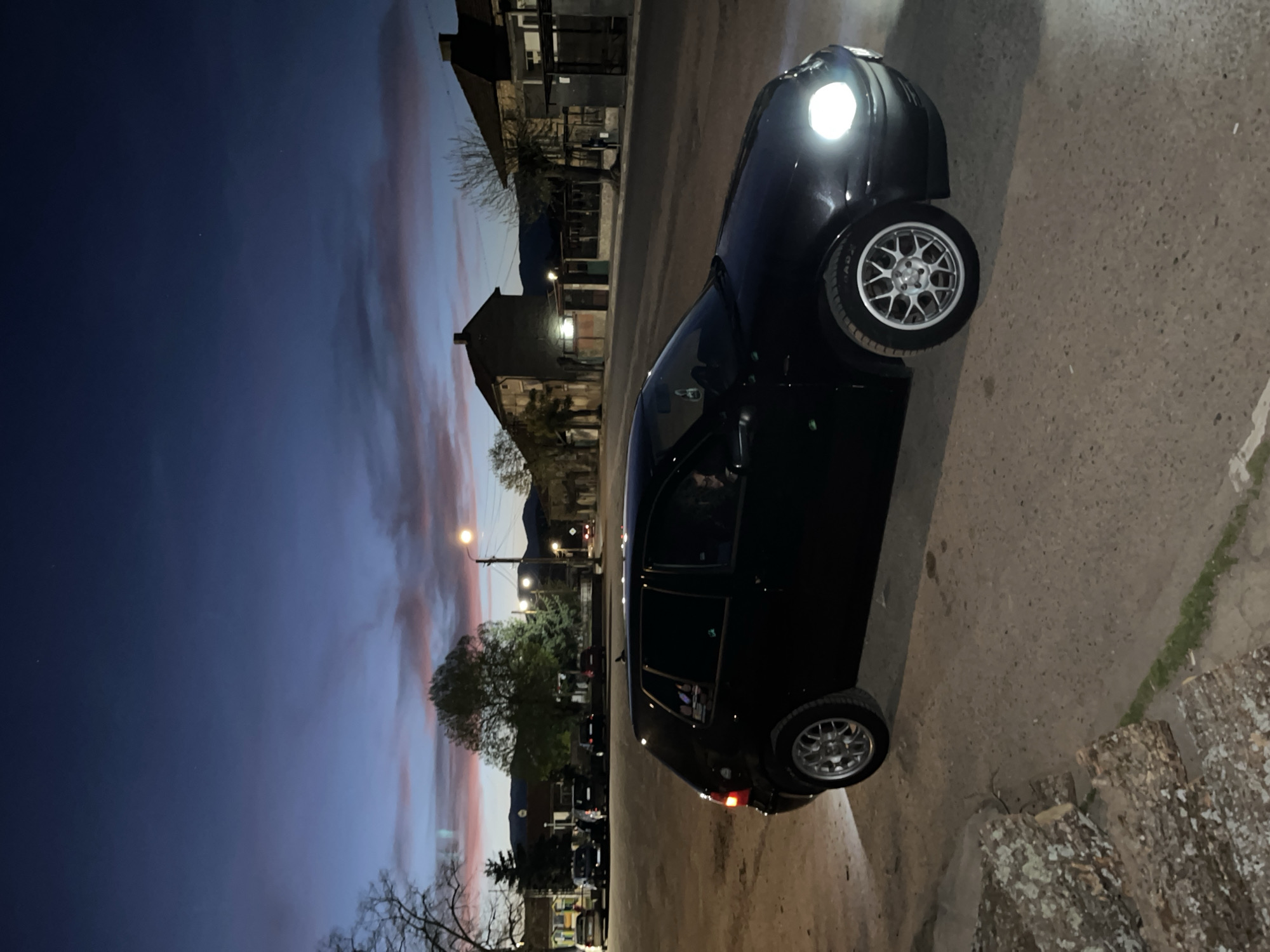 The image depicts a twilight scene on a street in Karavelovo, Bulgaria. A dark-colored (likely black) car, possibly a hatchback with bright headlights, is parked or driving on the right side of the frame. Its multi-spoke silver alloy wheels are prominent, and the word "BADXX" is faintly visible on the sidewall of the front tire. A reflection of a person's head can be seen in the car's side window.
The paved street, made of asphalt or concrete, shows textural details and slight wear. In the foreground on the right, a stack of cut logs, possibly for firewood, rests on the ground.
Buildings with traditional architecture line the street in the mid-ground and background, some appearing to be homes or small establishments. Illuminated streetlights cast warm light along the road, stretching into the distance. Bare and leafy trees are interspersed among the buildings. In the far background, distant mountains are silhouetted against the horizon.
The sky dominates the upper and left parts of the frame, displaying the distinct colors of dusk or twilight. It transitions from a deep, dark blue at the zenith to soft orange and pink streaks near the horizon, indicating a sunset or sunrise, though twilight is most likely given the artificial lights. The overall impression is one of peaceful serenity as day turns to night.