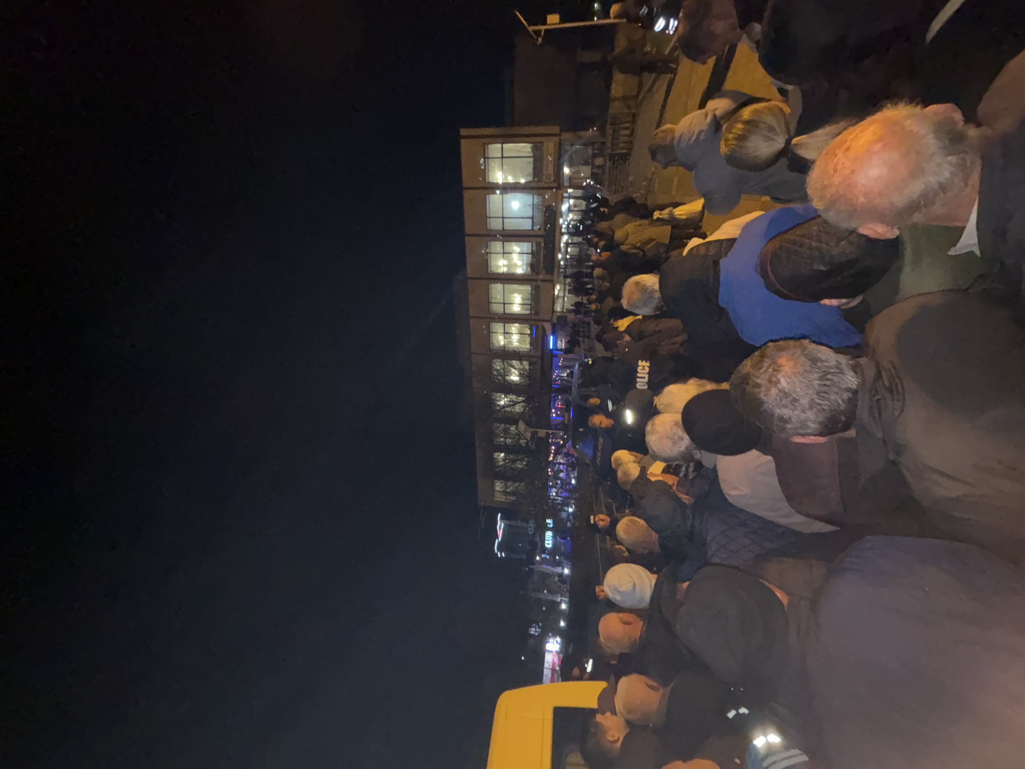 The image shows a large group of people gathered outside a building at night. The building appears to be a multi-story structure with illuminated windows. A uniformed police officer is visible in the crowd. The location is Kardzhali, Bulgaria.