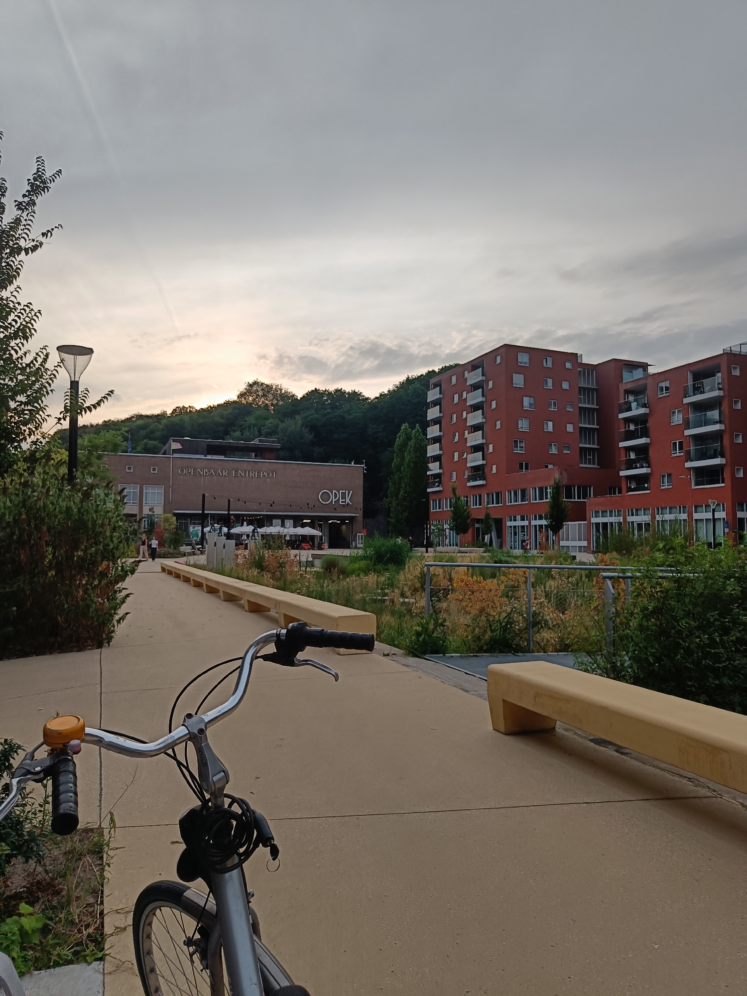 A silver bicycle is parked on a paved walkway in front of a building with the text "OPENBAAR ENTREPOT" and "OPEK" on its facade. The building is surrounded by greenery and trees. To the right of the building are modern red apartment buildings with balconies. The sky is overcast and appears to be dusk. A yellow bench is situated on the right side of the walkway. The scene is in a park-like setting within a city.