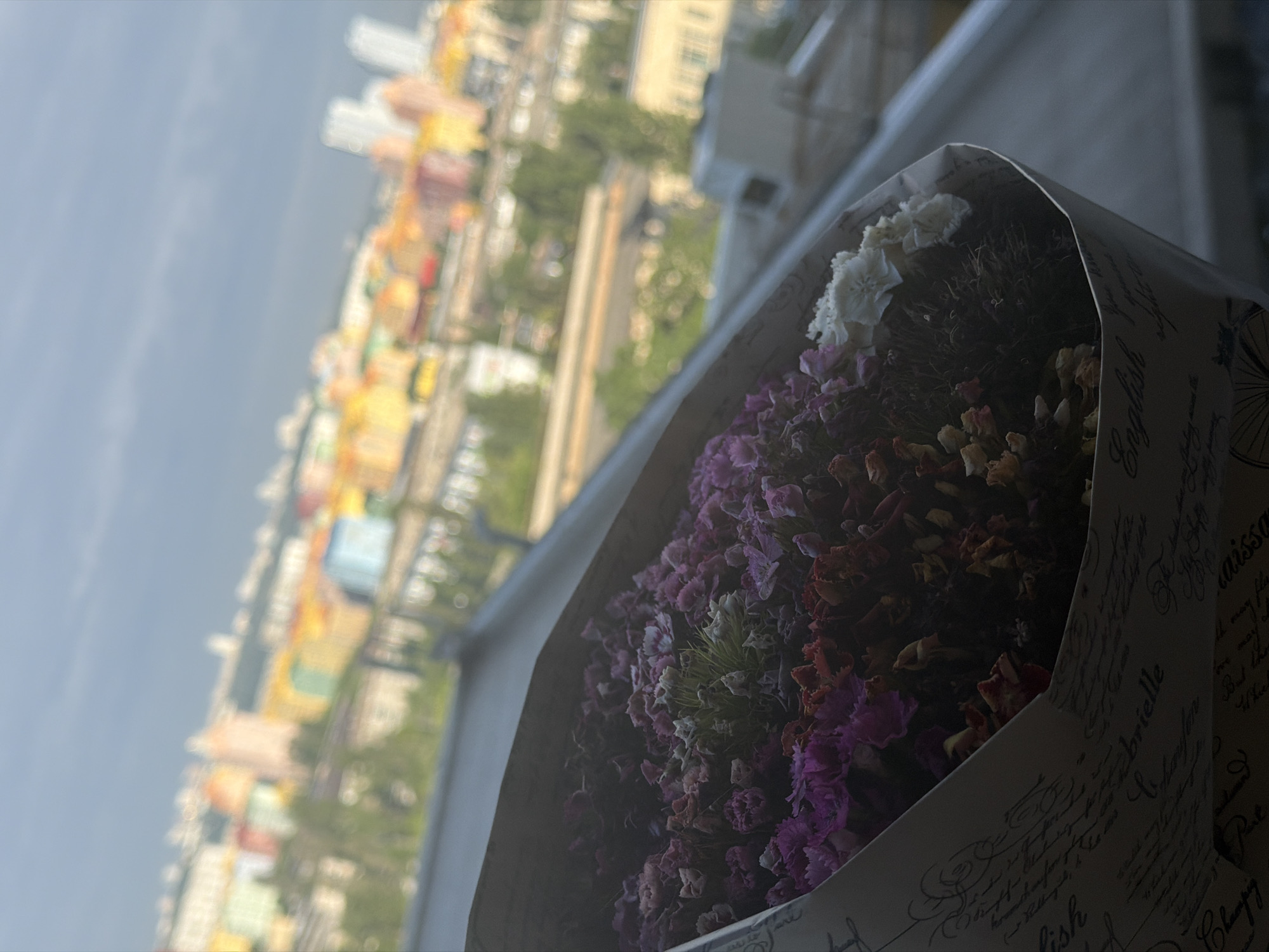 A bouquet of dried flowers is held in front of a blurred cityscape. The flowers are predominantly pink and white, with some dried brown and orange petals. The bouquet is wrapped in paper decorated with cursive writing. In the background, a city skyline is visible with buildings of various colors, including pink, yellow, blue, and orange. The sky is overcast with gray clouds. The scene appears to be a view from an elevated position, possibly a balcony or window, overlooking the city. The location is stated to be Kyiv, Ukraine.