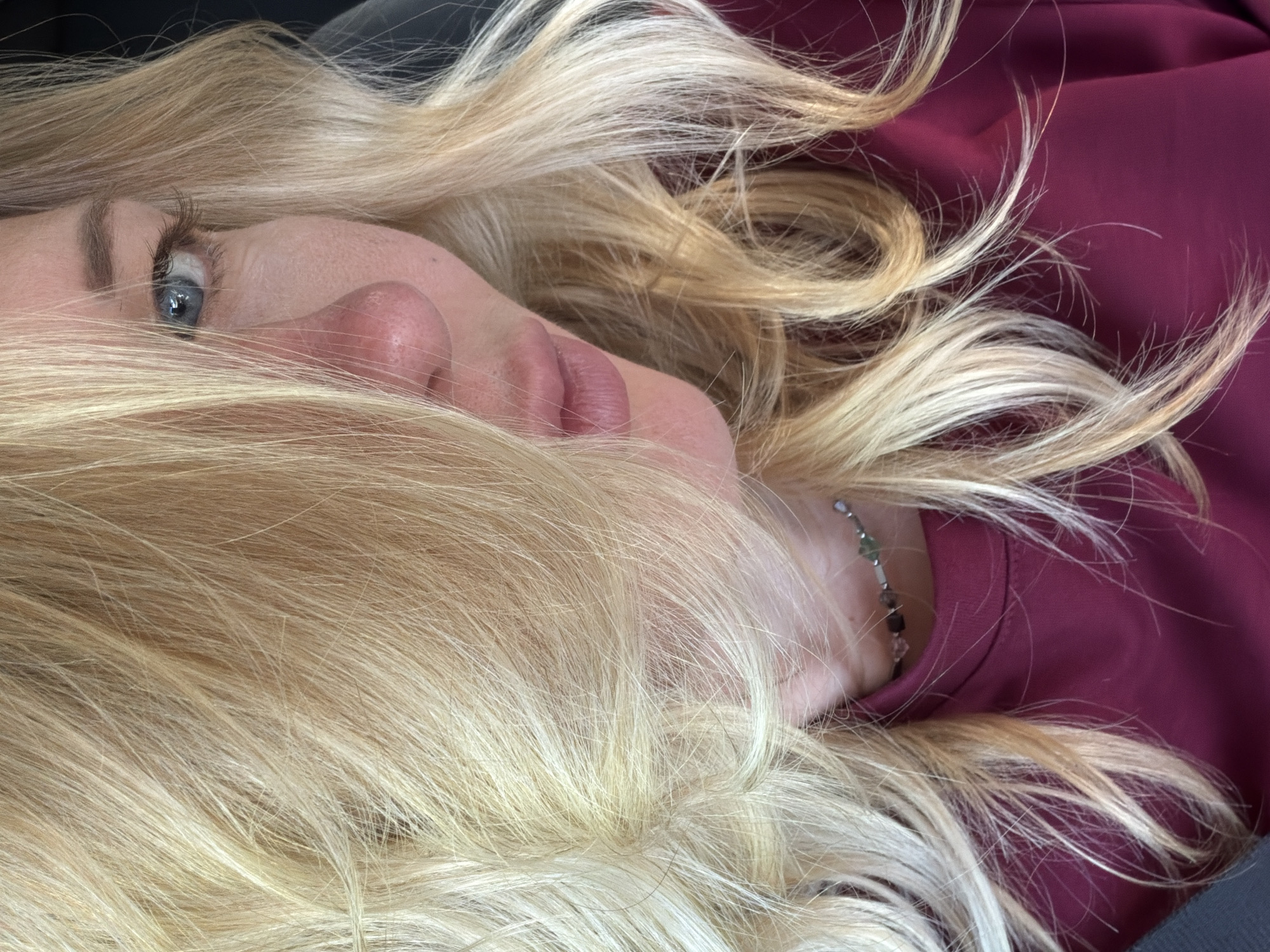 The image is a close-up, waist-up portrait of a woman with blonde hair, looking towards the right side of the frame. Her face is partially obscured by her hair, with her left eye visible. She is wearing a dark pink or magenta turtleneck shirt and a delicate beaded necklace. The background is out of focus, suggesting an indoor setting or a vehicle. There is no discernible location within Ново-Тебердинское сельское поселение, Russia.