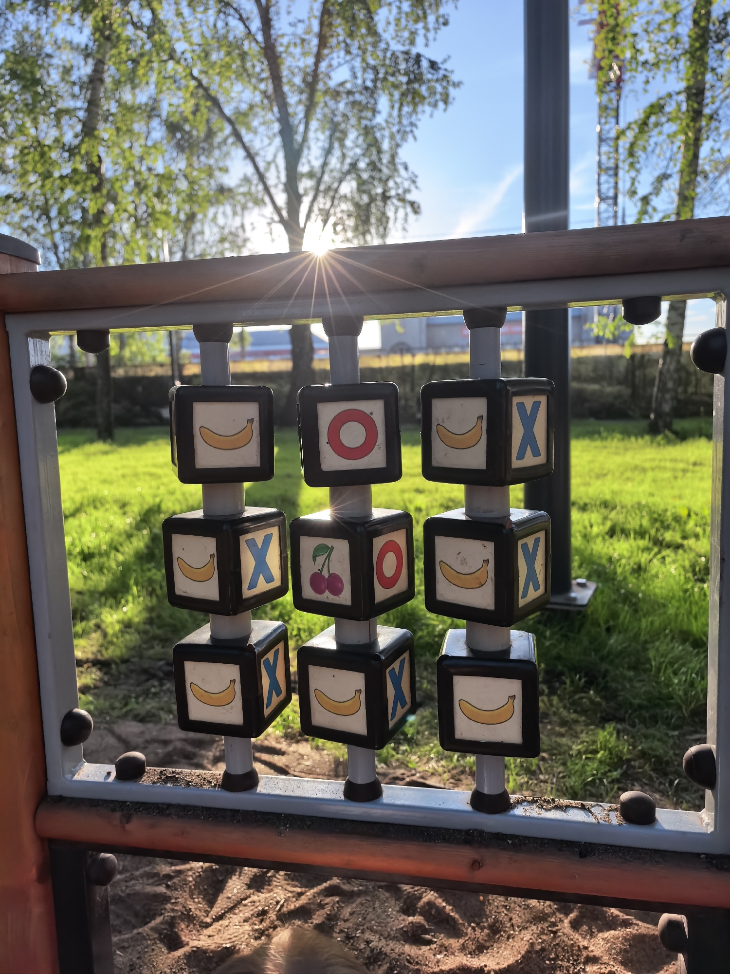 The image shows a Tic-Tac-Toe game on a playground, consisting of nine rotating cubes attached to vertical poles within a rectangular frame. Each cube has different symbols on its faces: bananas, X's, cherries, and O's. The cubes are arranged in a 3x3 grid. The frame is constructed of a combination of materials, including metal and possibly wood. The background includes green grass, trees, and a clear sky. The location is in the city of Sterlitamak, Russia.