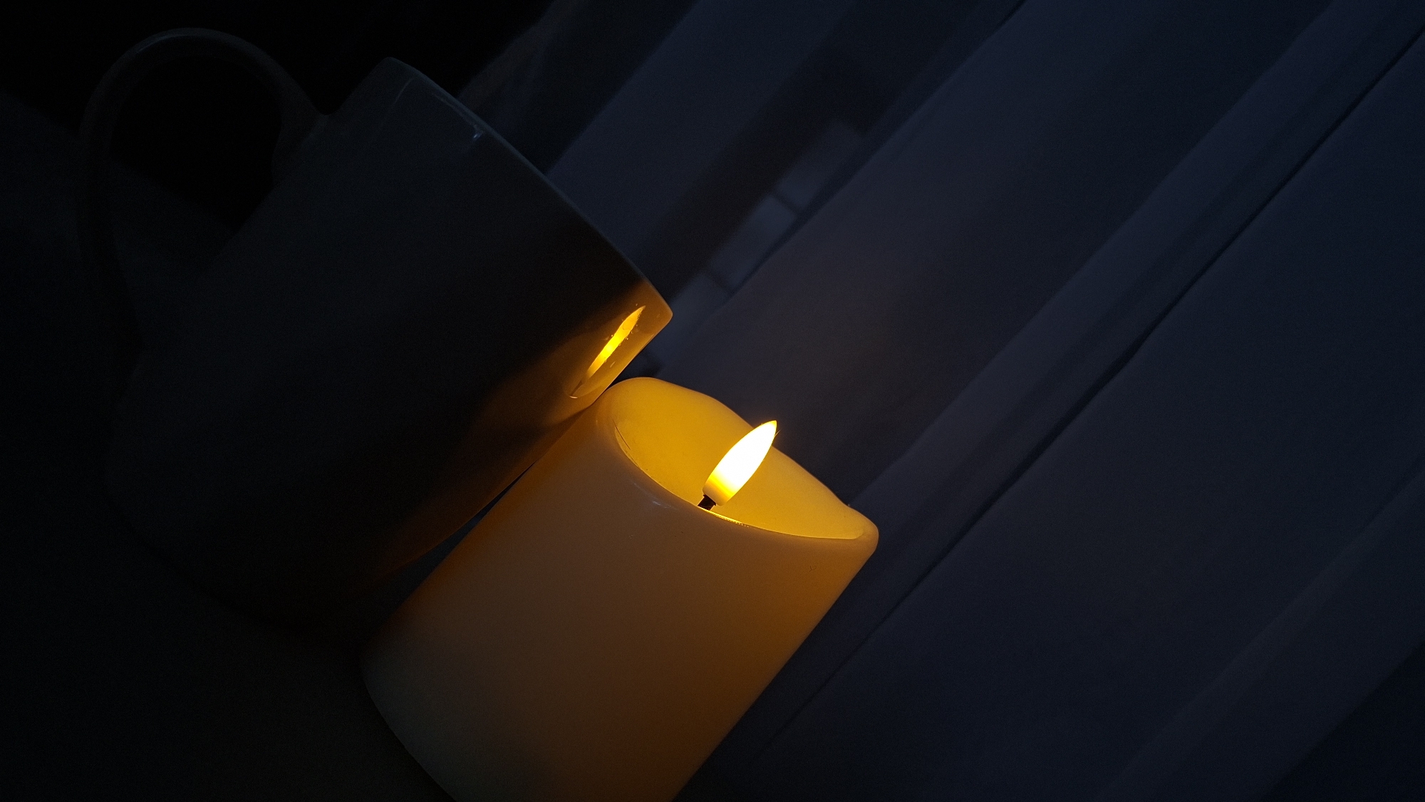 The image displays a close-up, low-angle shot of two lit candles placed near each other. The candles are cylindrical and appear to be made of a pale yellow material, possibly wax. Both candles emit a warm, yellow-orange light from their wicks, which are visible and appear to flicker. One candle is larger and more prominent, positioned towards the right side of the frame, with its flame casting a strong glow. The other candle is smaller and positioned to the left, partially obscured by the larger candle, with its flame also visible and contributing to the overall illumination. The background consists of dark, draped fabric, possibly curtains, that create a somber and intimate atmosphere. The lighting is dim, with the light from the candles being the primary source of illumination, highlighting the textures of the candles and the folds of the fabric. The image does not provide any specific geographical indicators or context that would allow for the identification of its location within the city of Myadzyel, Belarus.