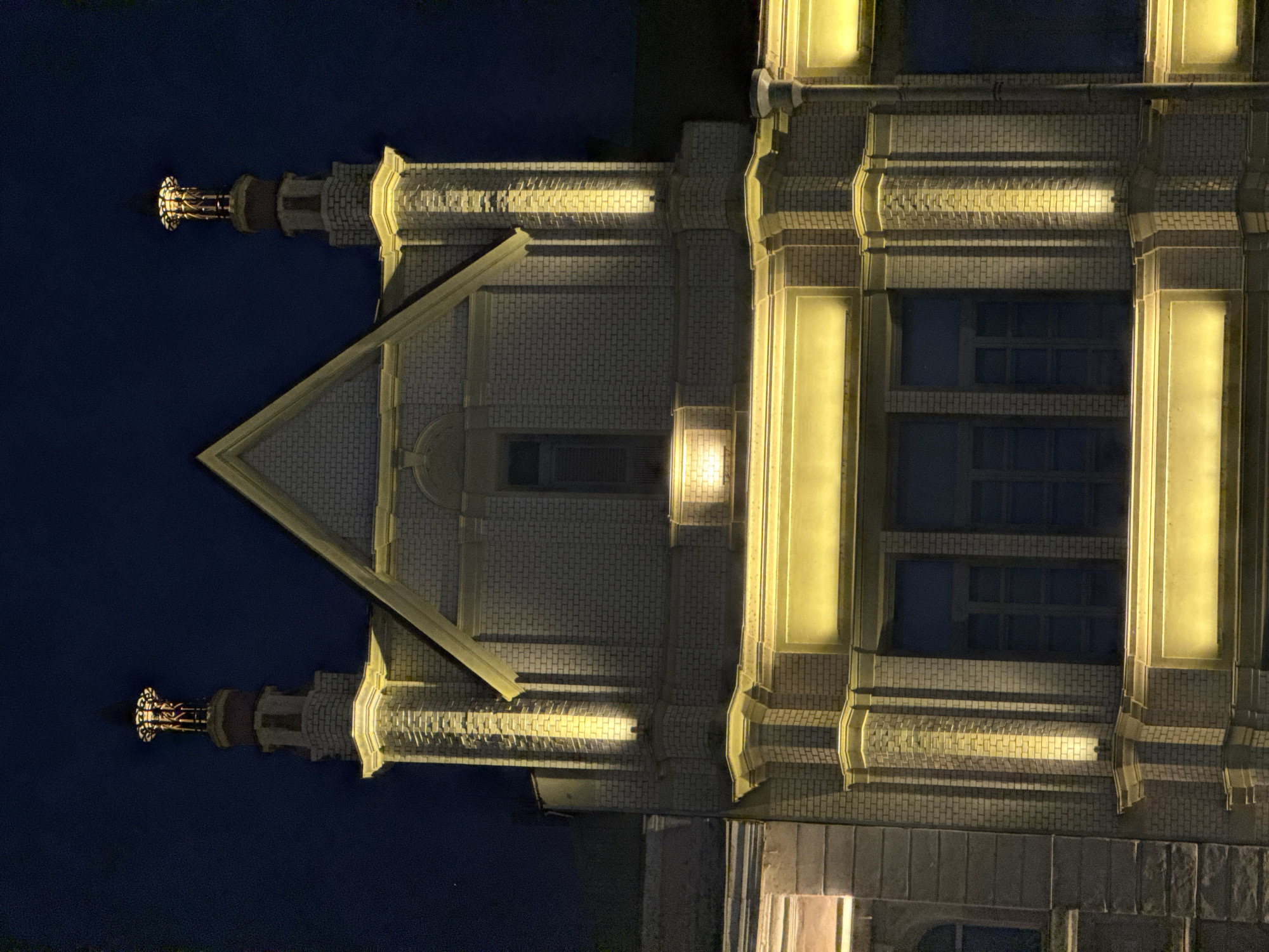 The image depicts the main building of the Nizhny Novgorod Fair in Nizhny Novgorod, Russia, captured at night. The structure is prominently illuminated by warm yellow artificial lights casting upwards from various points on the facade. The central section features a large, light-colored brick gable, beneath which is a recessed, dark rectangular window. Symmetrically flanking this central gable are two multi-tiered, square towers also constructed of light-colored brick. Each tower culminates in an ornate, dark metal finial or cupola with intricate detailing, which is also illuminated from within or below. The building's facade displays detailed brickwork, interspersed with decorative pilasters, cornices, and friezes, all highlighted by the artificial lighting. The windows throughout the visible portion of the building are dark, reflecting the deep blue-black night sky. No individuals, vehicles, or discernible actions are present in the scene.