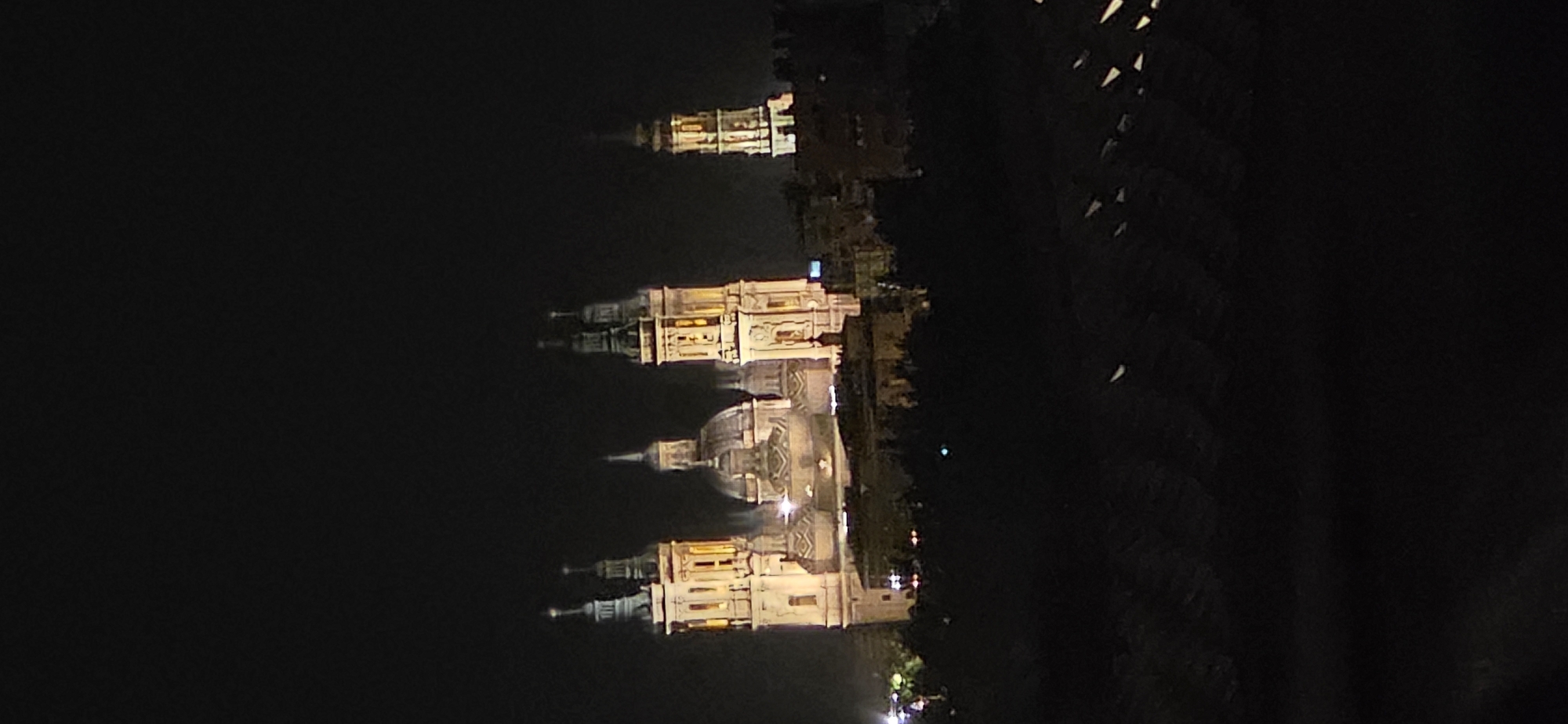 The image shows the Basilica of Our Lady of the Pillar (Basílica de Nuestra Señora del Pilar) in Zaragoza, Spain, at night. The illuminated facade of the basilica, with its prominent towers and domes, stands out against the dark sky. The foreground is obscured by darkness, with some lights visible in the distance. The basilica is a Roman Catholic church and a major pilgrimage site, known for its Baroque architecture.