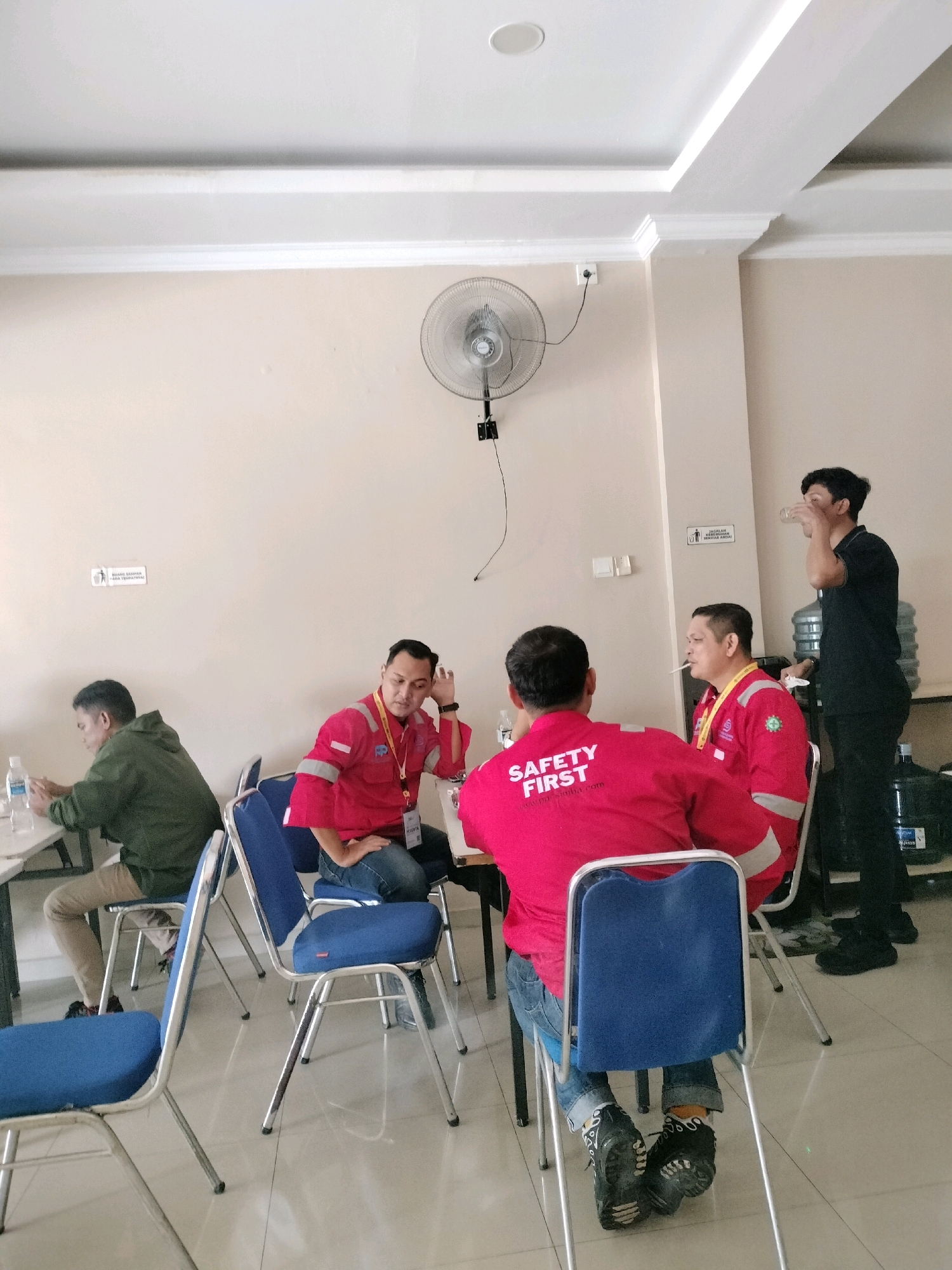 In Batam, Indonesia, five men are gathered indoors, near a wall with a ceiling fan. Two men in red shirts are seated, one with a cigarette in his mouth. Another man in a red shirt faces away from the camera, his back adorned with "SAFETY FIRST" printed in white. A fourth man, in a green hoodie, sits at a table with a water bottle and phone. The fifth man, in a black shirt, stands and drinks from a glass. Blue chairs and a water cooler are present. The scene appears to be a casual meeting or break area.