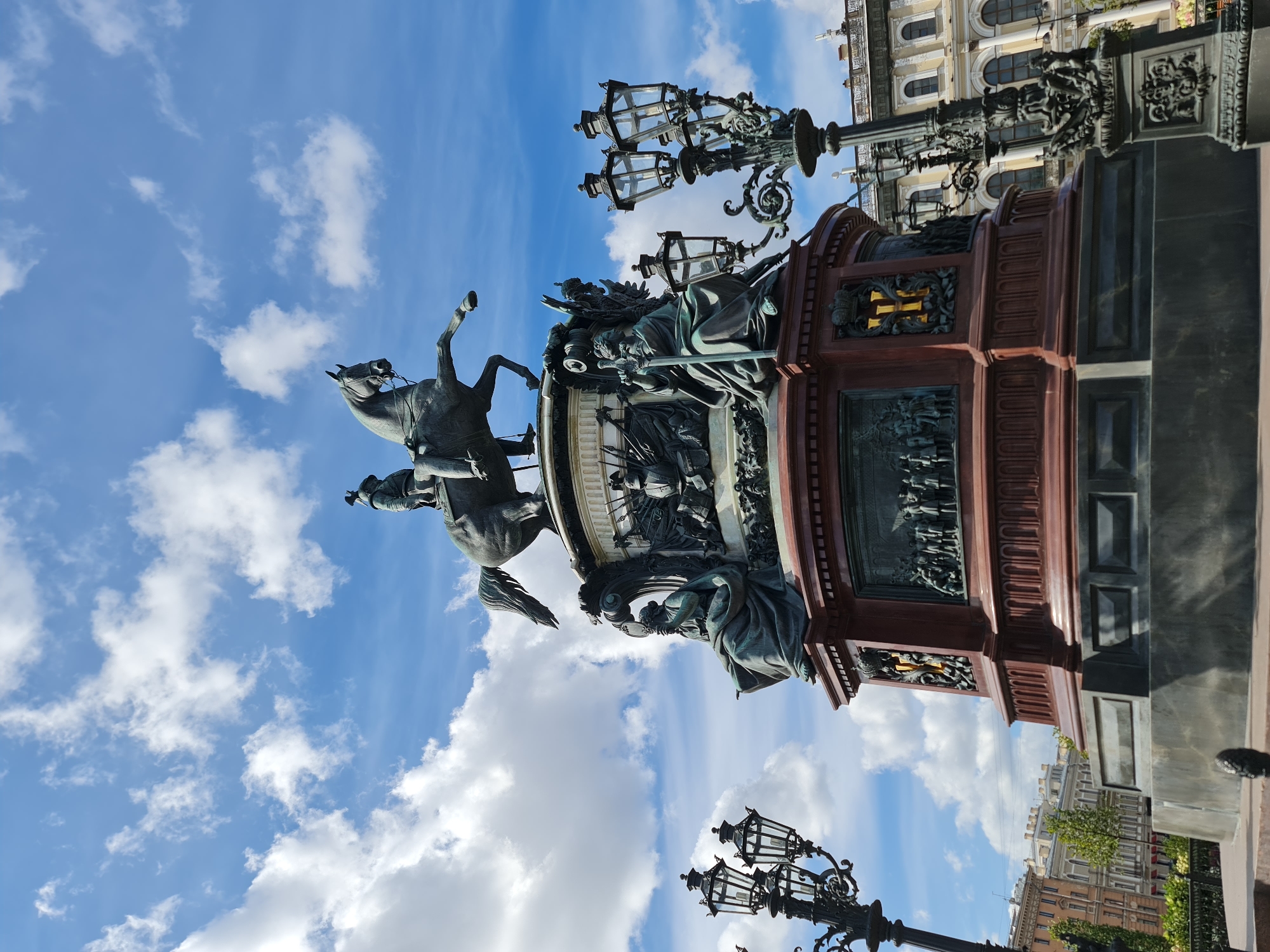 The image shows the Bronze Horseman, a monument to Peter the Great, located on Senate Square in Saint Petersburg, Russia. The monument features an equestrian statue of Peter the Great on a towering pedestal, with allegorical sculptures at its base. The statue is made of bronze, and the pedestal is granite. The monument is surrounded by ornate lampposts and is set against a backdrop of a blue sky with fluffy white clouds. In the distance, several classical-style buildings can be seen. The perspective is from below, looking up at the monument.
