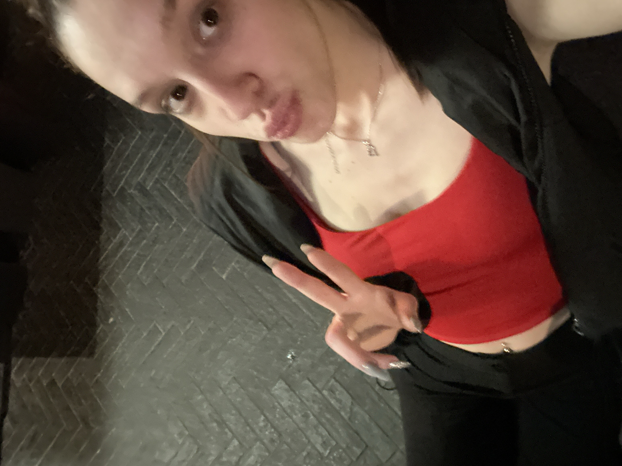 A young woman is posing for a selfie. She has light skin, dark hair pulled back from her face, and a silver necklace. She is wearing a red top and a black jacket, and black pants. She has a navel piercing. She is making a peace sign with her hand. The background is a dark herringbone brick pattern. It is not possible to determine the location as Varna, Bulgaria with the given information.