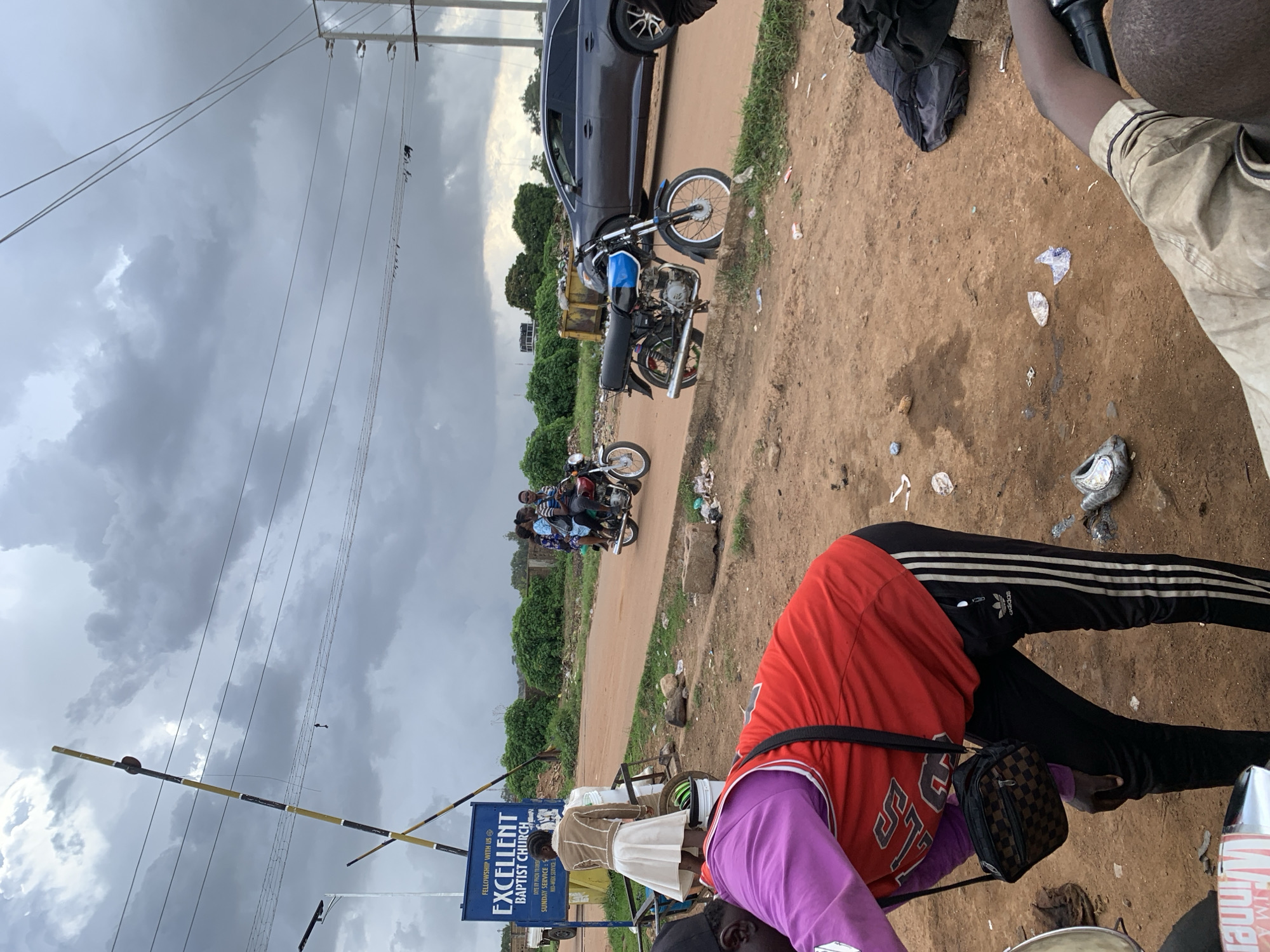 In Barakin Lerawi, Nigeria, a dusty, unpaved road is visible under a cloudy sky. To the left, a yellow and black striped barrier stands by a blue sign that reads "FELLOWSHIP WITH US EXCELLENT BAPTIST CHURCH." A woman in a brown jacket and white skirt stands near a water dispenser. In the middle ground, a motorcycle carrying three people rides down the road. To the right, a dark grey SUV is parked. In the foreground, a person wearing a red jersey and black Adidas track pants with a small checkered bag slung across their body is leaning down. Another person's arm in a light brown shirt is visible on the far right, seemingly holding onto a motorcycle handlebar. The ground is littered with various debris, including plastic bottles and wrappers.