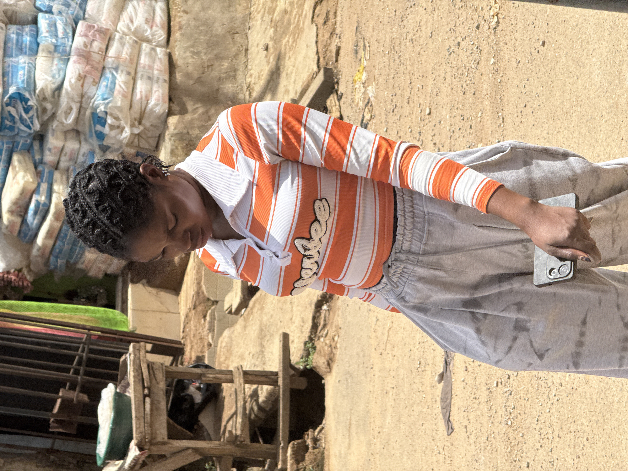 A young woman stands on a dirt surface in Abuja, Nigeria, holding a black smartphone in her right hand. She wears a long-sleeved, horizontally striped shirt with orange and white bands and light grey sweatpants. Her hair is styled in braids. Behind her, a pile of wrapped goods rests against a wall, alongside a deteriorated wooden structure. The ground is sandy and dry.