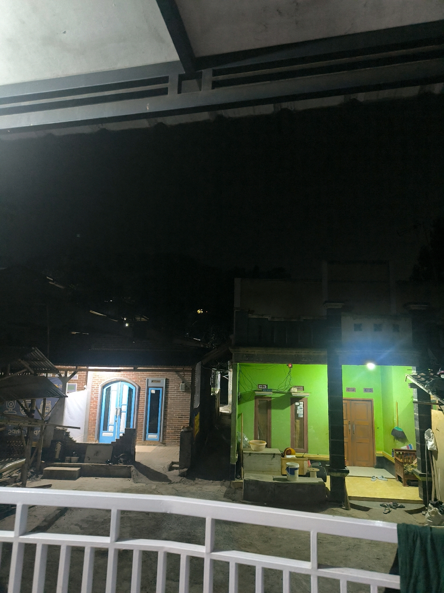 This image depicts a nighttime scene of two adjacent residential buildings in Cilengkrang, Indonesia. The building on the left is constructed with brick and features a light-colored wall with two windows. It has a prominent arched doorway with double doors painted blue, flanked by a blue-framed window on the right. In front of this building, there are two steps leading up to the entrance, and a small covered structure with a corrugated metal roof is visible to the far left, partially obscuring the building.
The building on the right is painted a vibrant green and appears to be a single-story structure with a wooden door. There are two windows on the green facade. A light illuminates the interior of this building, revealing some objects inside, including a wooden bench. The area in front of this green building is paved, and several pairs of flip-flops are scattered on the ground.
Both buildings appear to be residential houses. The scene is illuminated by artificial lights, with a bright light source in the green building and lighting from inside the brick building. The sky is dark, indicating it is night. In the foreground, a white metal railing is visible, suggesting a balcony or walkway from which the photograph was taken. The ground in front of the buildings is uneven and appears to be made of dirt or gravel. The overall impression is that of a humble residential area at night.
