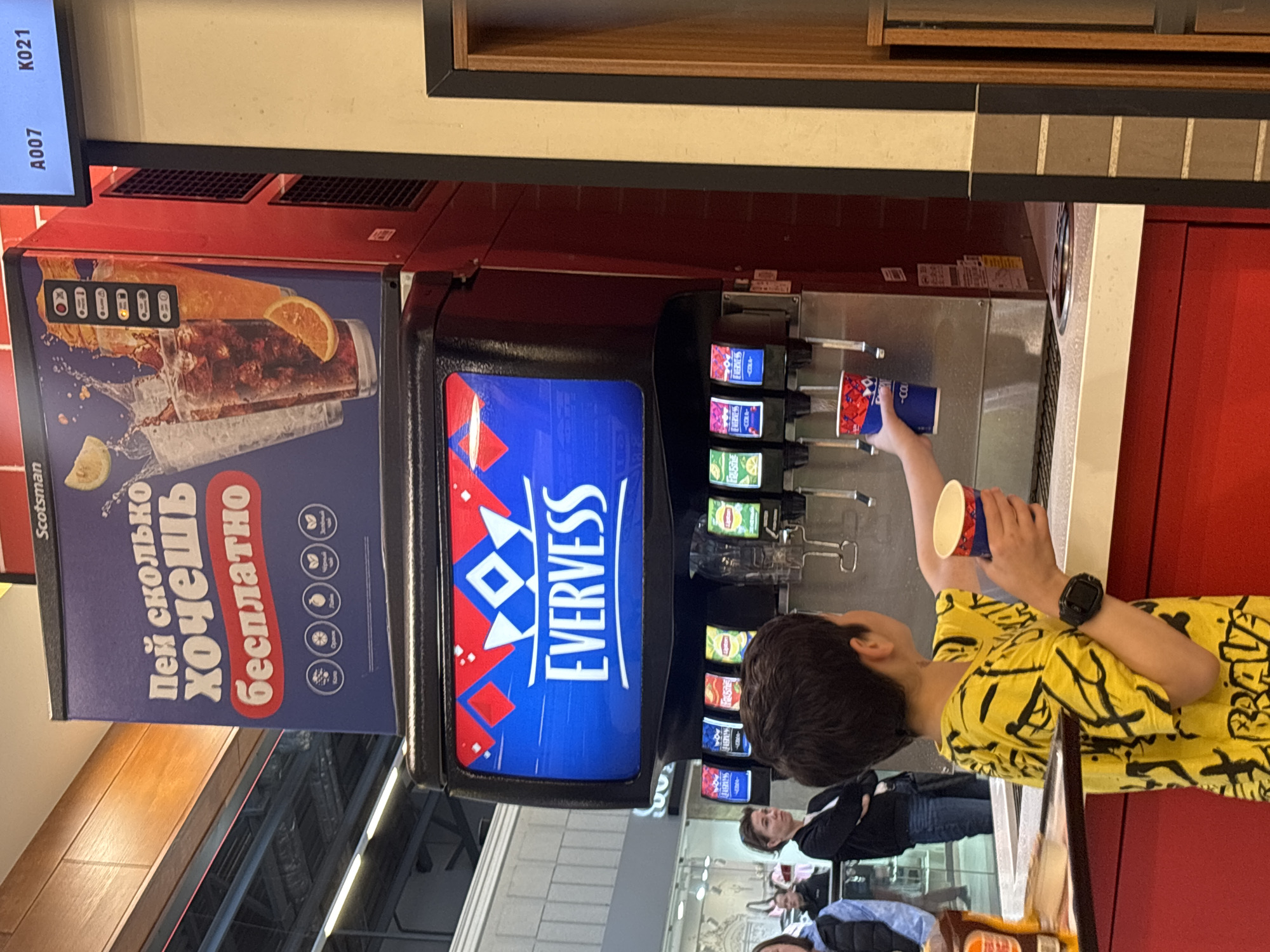 A medium shot, taken indoors, features a child filling a cup with soda from a soda fountain. The child, who appears to be a young boy, is wearing a bright yellow shirt with a black scribble pattern. He is holding a blue and red patterned cup up to the fountain, and a black smartwatch is visible on his left wrist.
The soda fountain is a commercial model, with branding that includes the word "EVERVESS" in large white letters on a blue background. To the left of the fountain is a panel with a graphic of a soda drink and text in Russian, including the word "бесплатно" (free).
In the background, other individuals are present, though they are not the primary focus of the image. They appear to be customers in what seems to be a casual dining or food court setting. The environment suggests a public place, likely a restaurant or food court, possibly within a shopping mall. The location is in the city of Unknown, Unknown.