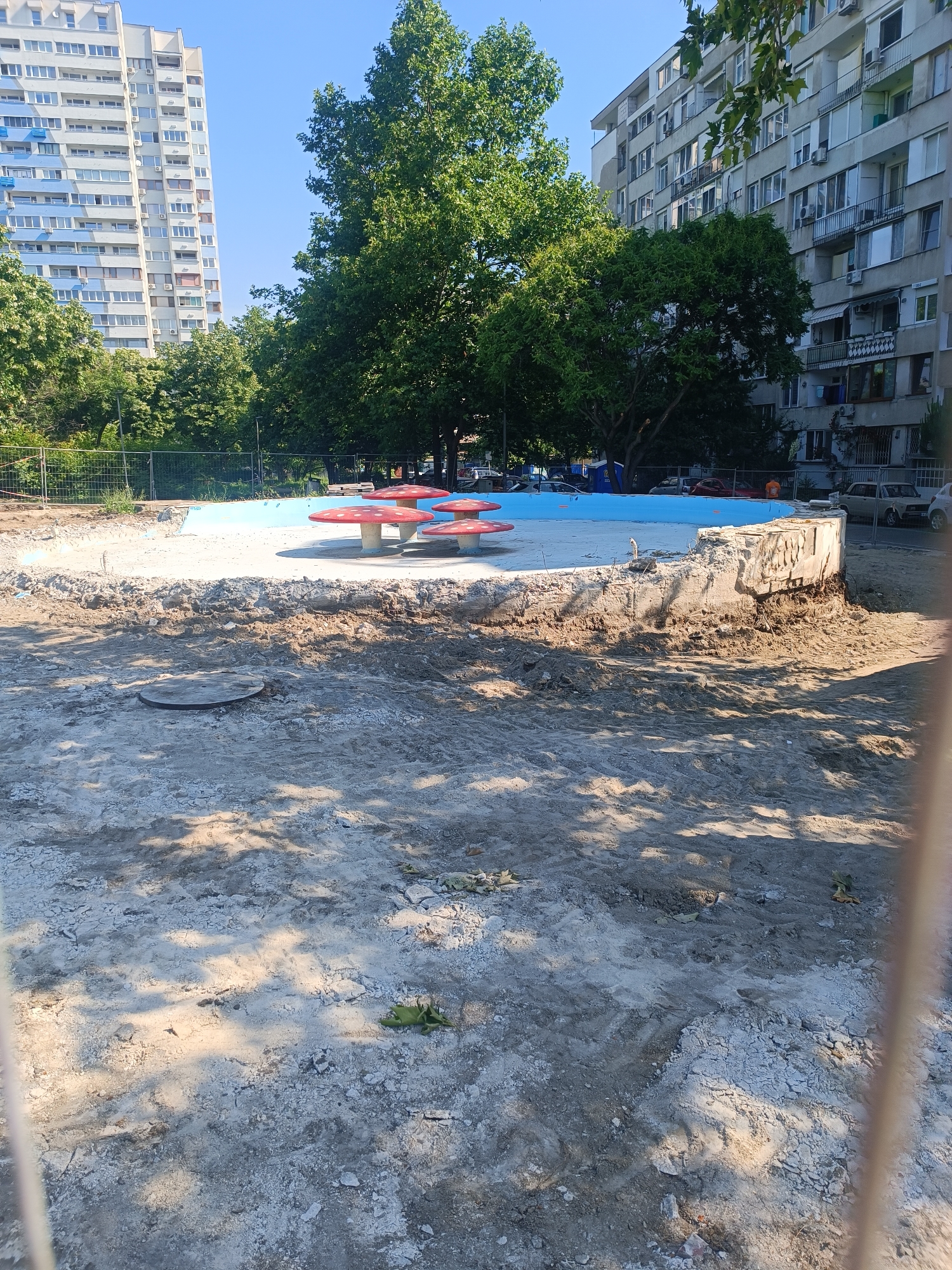 The image depicts an outdoor scene in an urban environment, likely a park or a public space undergoing renovation. In the foreground, the ground is uneven, covered with sand, dirt, and debris, suggesting construction or demolition work. A circular metal cover, possibly a manhole, is visible on the sandy ground.
In the midground, a circular area with a light blue surface is partially visible, bordered by a rough, concrete structure that appears to be a wall or a raised edge. Several red and white mushroom-shaped decorative elements are arranged on this blue surface, suggesting a children's play area or a decorative fountain.
In the background, a tall, multi-story apartment building with a light blue and white facade stands to the left, its windows and balconies clearly visible. To the right, another apartment building, gray in color, with numerous windows and balconies, dominates the background. Green trees with dense foliage are scattered throughout the scene, providing shade and a contrast to the urban structures. A metal fence is visible in the far background, bordering the area. The sky is clear and blue.
While the image is not definitively identifiable as being in Burgas, Bulgaria, the architectural style of the apartment buildings and the general urban setting are consistent with typical Bulgarian cityscapes. The presence of a construction or renovation site, along with the decorative elements, suggests a public space being revitalized. There are no visible people in the image. The overall impression is one of a public space in transition.