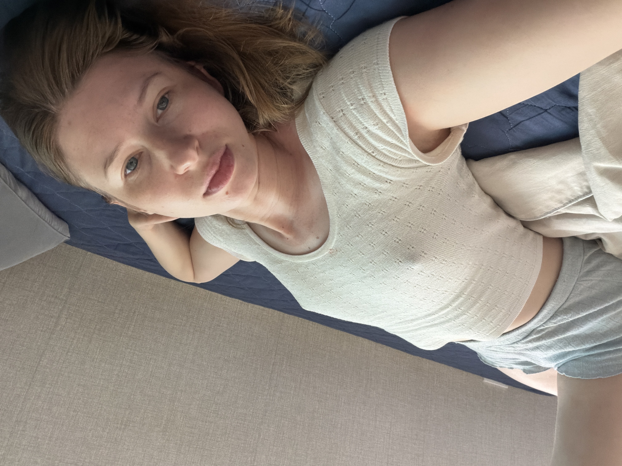 A young woman lies on a bed with a blue quilted comforter in a room with a textured beige wall in the background. She is wearing a white ribbed tank top and light blue shorts. Her head is tilted back, and her eyes are looking towards the camera. Her hair is brown and spread out on the comforter. The scene is lit by overhead lighting. The overall impression is a casual and relaxed pose.