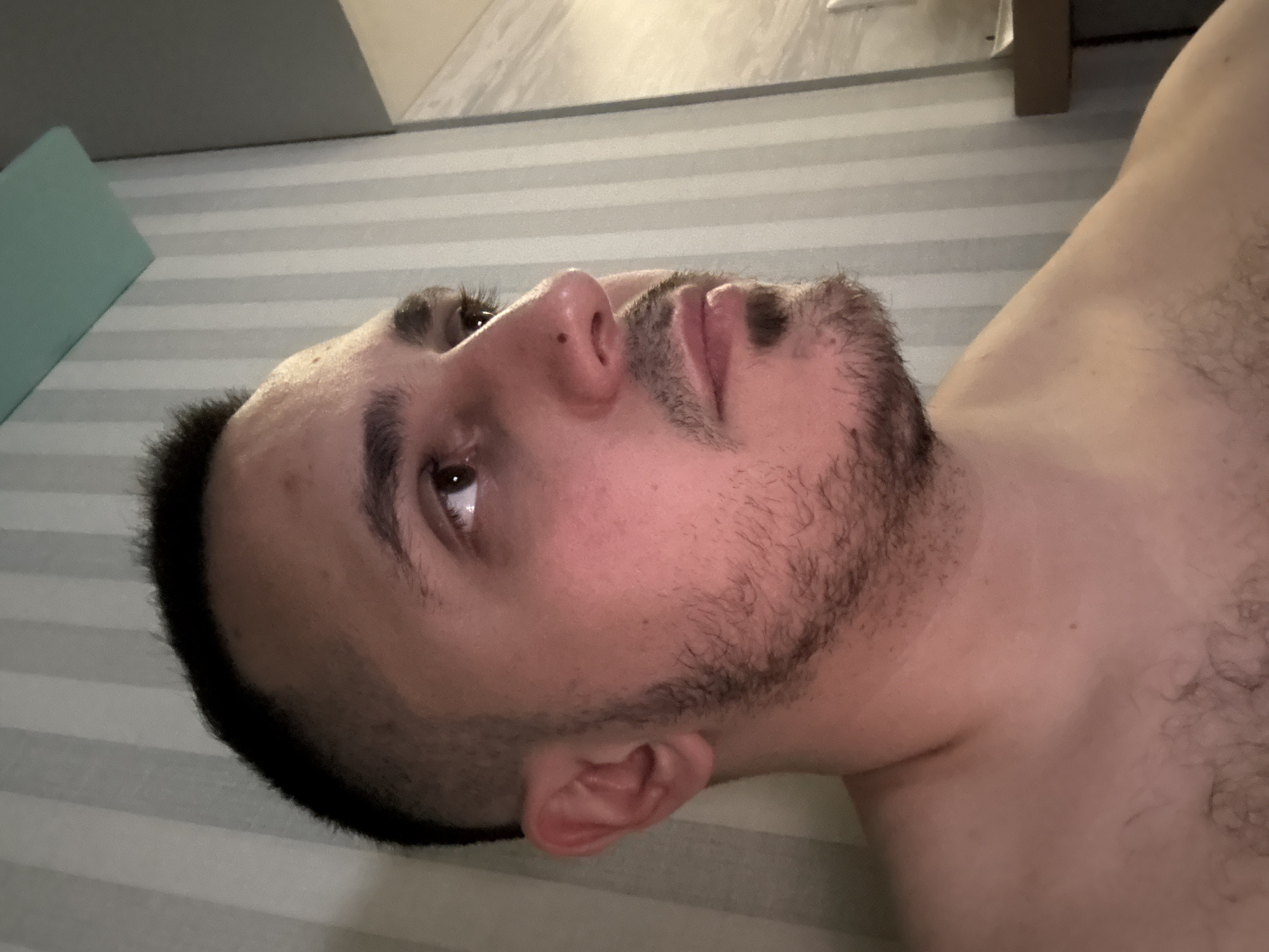 A close-up, horizontally oriented image captures the side profile of a person, likely male, lying down. The subject's head is positioned towards the left of the frame, with their face angled upwards and towards the right. They have dark, short hair and a short, dark beard or stubble covering their jawline and chin. Their skin appears fair, with some redness visible on the cheek. The person's left eye is open, gazing upwards. Their nose, mouth, and ear are clearly visible.
The subject is shirtless, revealing their right shoulder and upper chest, which has some body hair. The background consists of a light-colored surface with horizontal stripes, possibly a bedsheet or a patterned wall, suggesting an indoor setting. In the upper left corner of the background, a light blue or teal object is partially visible. The lighting appears to be artificial, casting soft shadows, and no visible text is present. The person's expression is neutral, conveying a calm or contemplative mood.