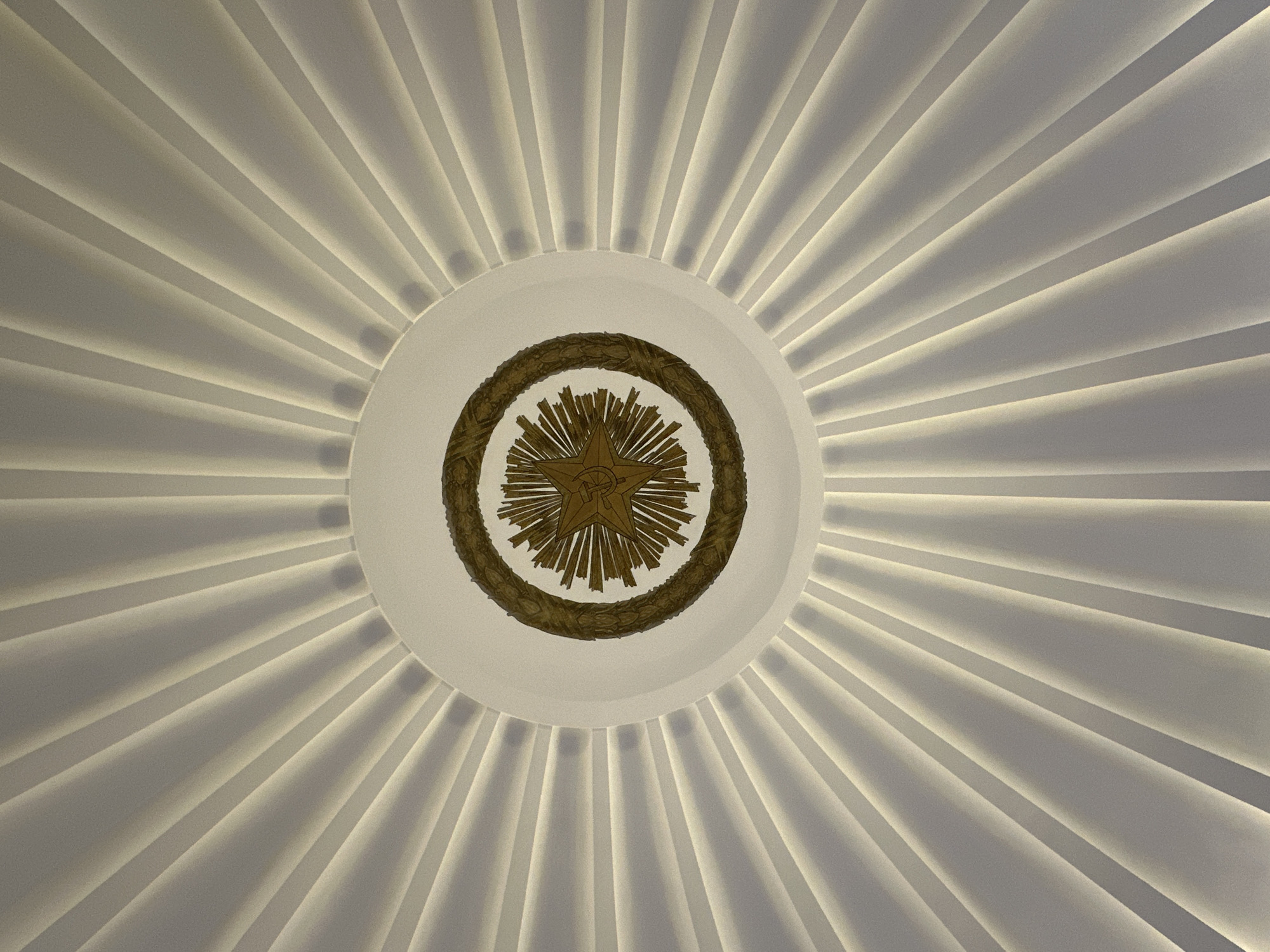 The image shows a close-up, upward view of a ceiling featuring a star emblem surrounded by a wreath. The ceiling itself is designed with radiating lines, creating a dramatic, sunburst-like effect with integrated lighting that casts soft shadows. The star emblem is gold-colored and depicts a hammer and sickle, a symbol associated with the Soviet Union. The wreath appears to be made of laurel leaves. The overall aesthetic is grand and possibly institutional. This emblem and ceiling design are characteristic of the Moscow Metro system, specifically found in some of its older and more ornate stations, such as the Mayakovskaya station, which is known for its Art Deco style and Soviet iconography.