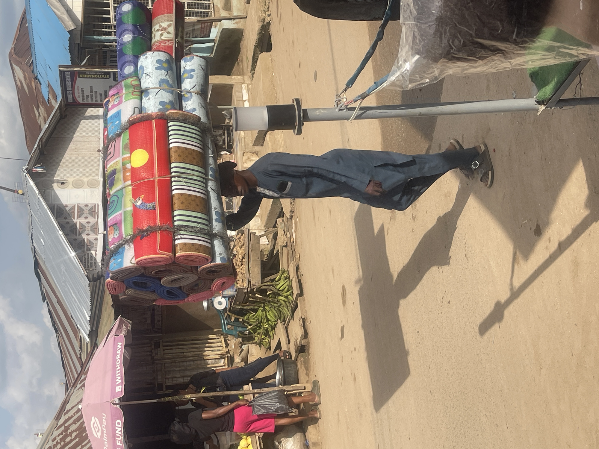 A man in a blue traditional outfit is walking on a dirt road, carrying a large stack of rolled-up mats on his head. The mats are colorful and patterned. He is wearing sandals. In the background, there are buildings with corrugated iron roofs, stalls, and a pink umbrella with "PalmPay FUND" and "WITHDRAW" written on it. Some people are standing near the stalls, and there are bunches of bananas and other produce displayed. The scene appears to be a market in Abuja, Nigeria, under a cloudy sky.