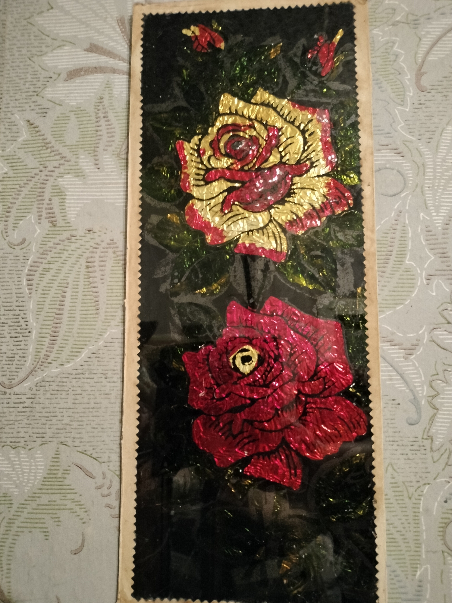 The image displays a vertical decorative item featuring two stylized roses made of foil and glitter against a black background. The top rose is gold with red accents, and the bottom rose is entirely red with a yellow center. The item has a sawtooth edge on the right and is mounted on a piece of cream-colored cardstock. The background to the left of the item is a light-colored surface with a repeating pattern of green floral motifs and faint horizontal lines. There are no people or identifiable landmarks in the image. The location of Lykhove, Ukraine, is not depicted or suggested by the image content.