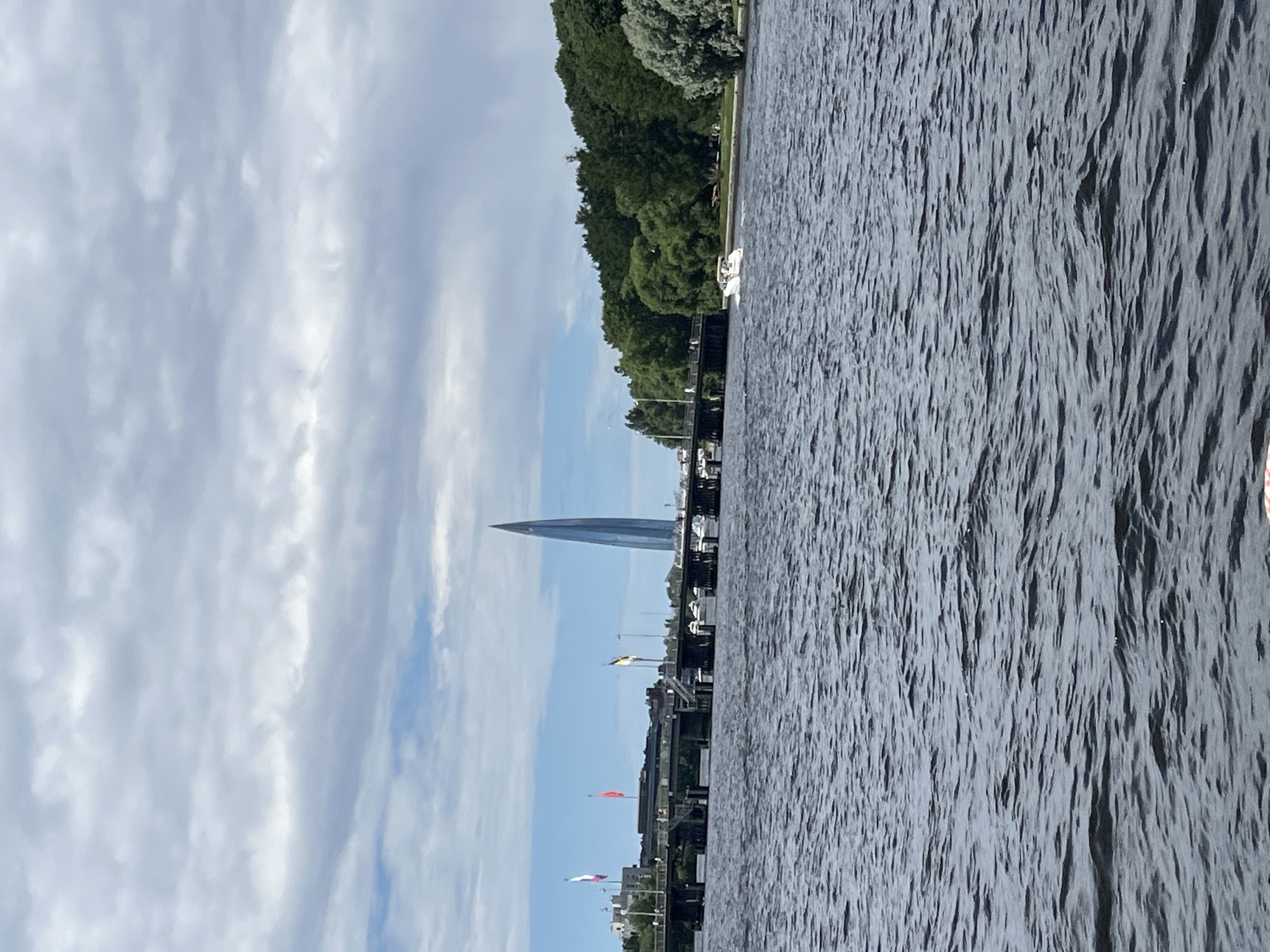 The image depicts a wide, dark body of water, likely a river or canal in Saint Petersburg, Russia, with gentle waves rippling across its surface. In the background, a modern, glass-covered skyscraper with a distinctive pointed spire dominates the skyline. This is the Lakhta Center, located in the Primorsky District of Saint Petersburg.
A long, elevated bridge stretches across the water, supporting vehicular and pedestrian traffic. Several flags, including the Russian tricolor, are visible on the bridge. A small white motorboat with two people on board is moving across the water towards the right side of the frame. The far bank of the water features a dense line of lush green trees, suggesting a park or natural area. The sky above is partly cloudy, with patches of blue sky peeking through a layer of grey clouds. The overall scene conveys a sense of urban development integrated with natural elements.