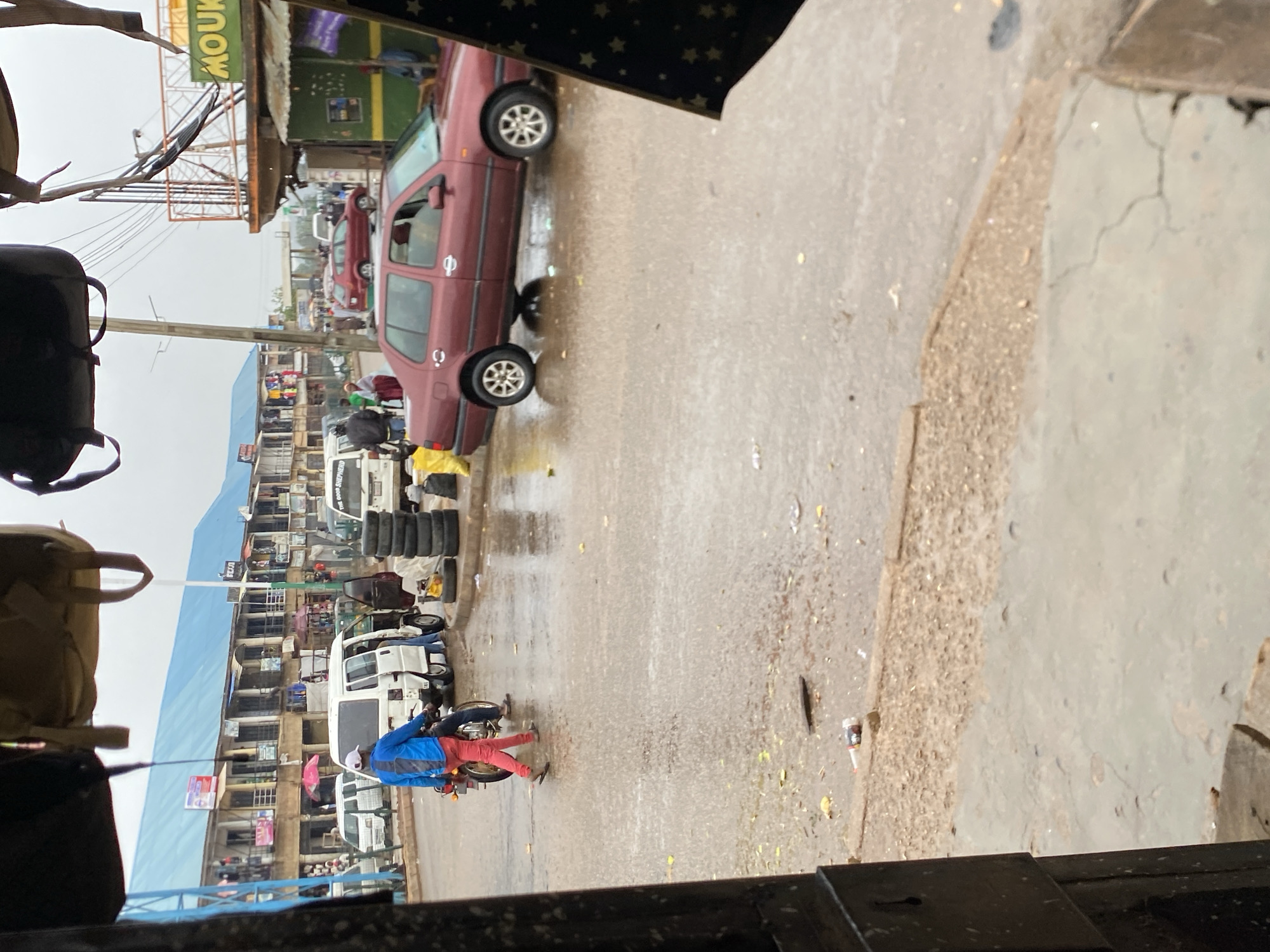 The image shows a street scene in Bukuru, Nigeria, on a rainy day. A red car is parked on the side of the road. Several people are present, including a man on a motorcycle wearing a blue jacket and red pants. Buildings line the street, with businesses and shops visible. Tires are stacked near a white van. The street surface is wet, and there is debris scattered around.