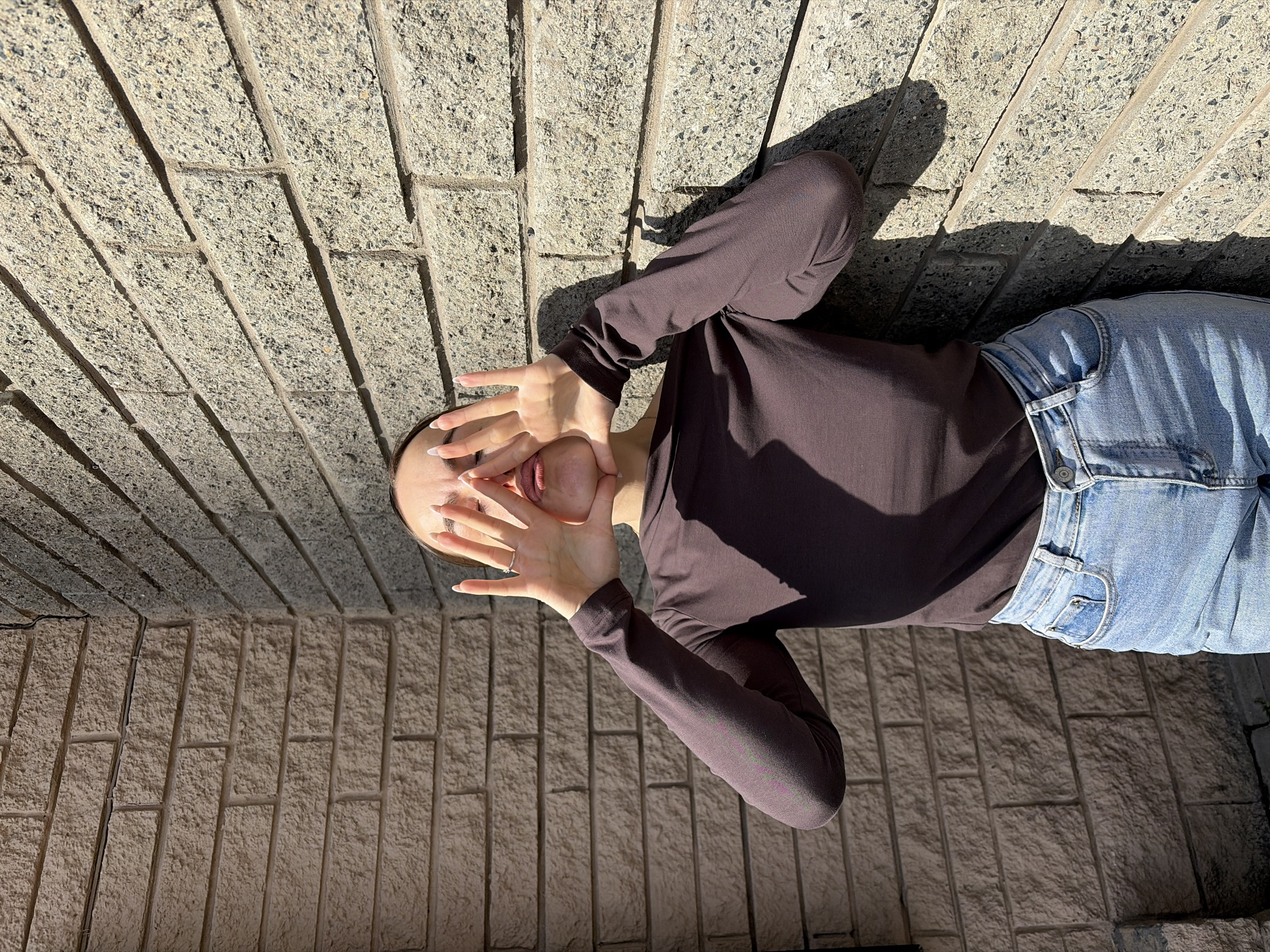A woman is positioned against a brick wall backdrop. She is wearing blue jeans and a long-sleeved brown shirt. Her hands are raised, partially covering her face. The setting appears to be outdoors, with sunlight casting shadows on the wall and the subject. No evidence suggests the location is Izhevsk, Russia.