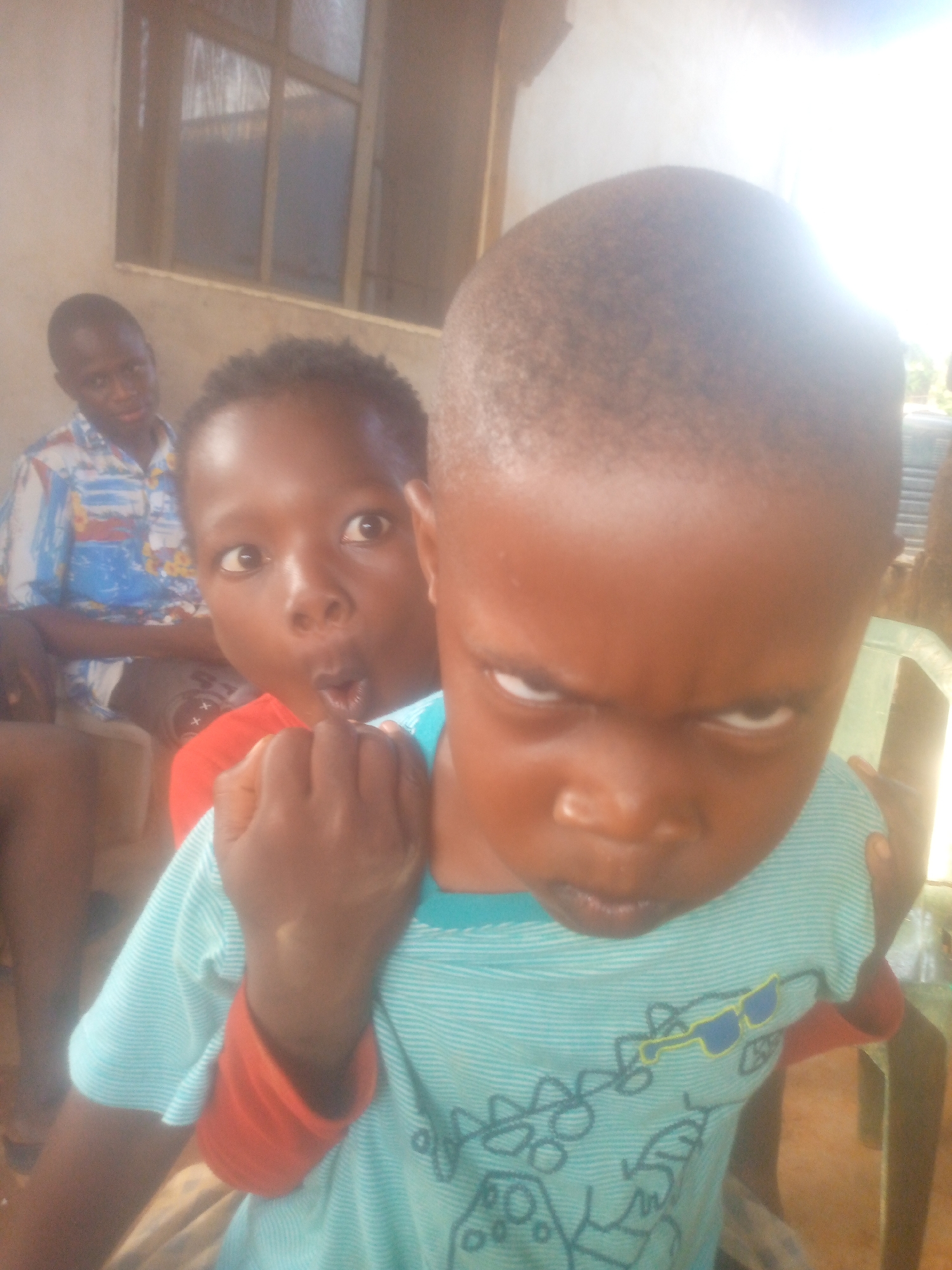 The image shows two children and an adult man in an indoor setting, possibly in the city of Unknown, Unknown. The primary focus is on the two children, with one child in the foreground displaying a serious expression and furrowed brow. He wears a light blue shirt with a design on it. Behind him, another child is visible, wearing a red shirt and making a surprised facial expression. An adult man sits in the background, slightly out of focus and wearing a patterned shirt. A window is visible in the upper left corner, and a chair appears on the right side of the frame.
