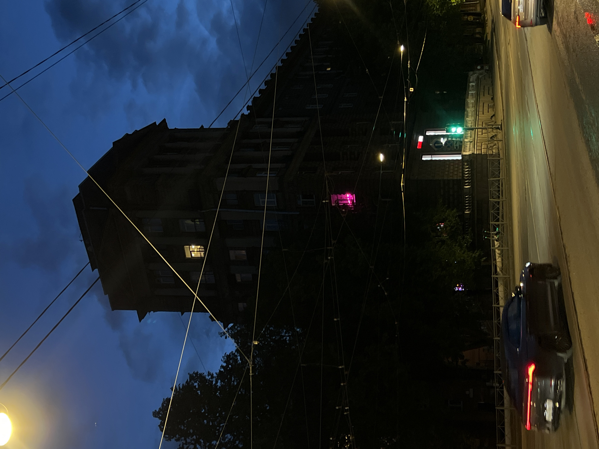 An exterior, low-angle, vertical, eye-level shot captures a dark, imposing apartment building at dusk on a street in Saint Petersburg, Russia. The building, characterized by its classical architectural style with visible columns and ornate detailing, is silhouetted against a deep blue, cloudy sky. A single window on the upper floors emits a warm, yellow light, suggesting occupancy. Further down the facade, a window glows with a vibrant pink light. The foreground is dominated by a complex network of dark overhead wires, likely for a tram or trolleybus system, crisscrossing the frame. A dark-colored luxury sedan is captured in motion on the asphalt road, its taillights illuminated. To the right, a traffic light displays a green signal, indicating traffic flow. The scene is illuminated by streetlights, casting a warm glow on the road surface, and the overall atmosphere is one of twilight transitioning into night.
