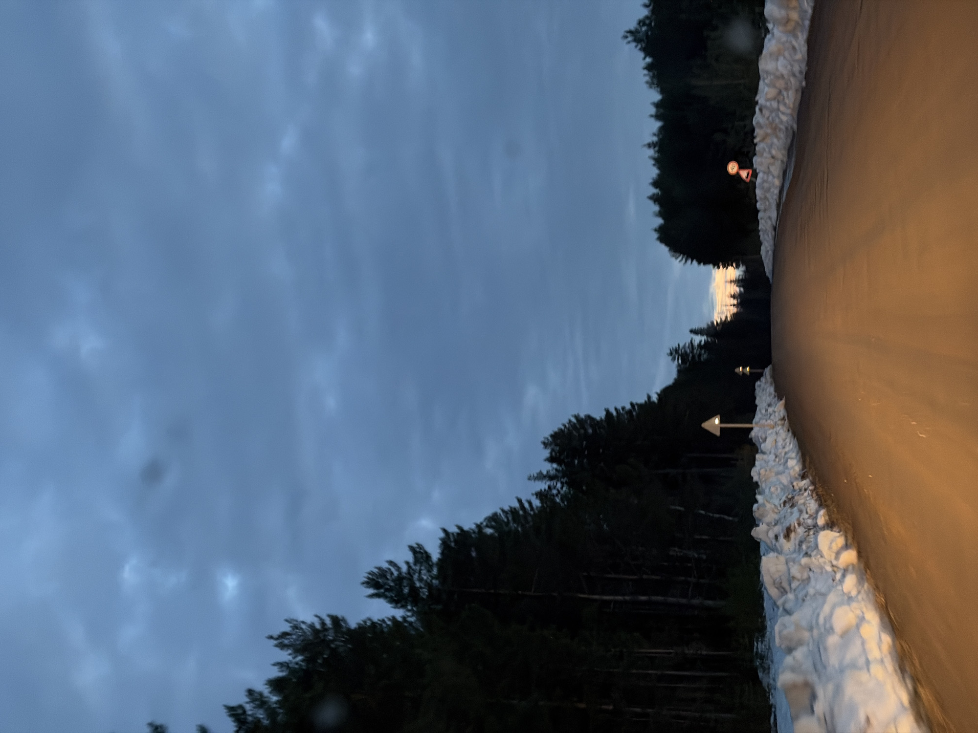 The image depicts a road in a forested area during twilight. The sky is overcast. The road surface is light brown. The sides of the road are lined with snow. A traffic sign with a white arrow pointing left is visible on the left side of the road. Another traffic sign is visible further down the road on the left. The location is Kostovaty, Russia.