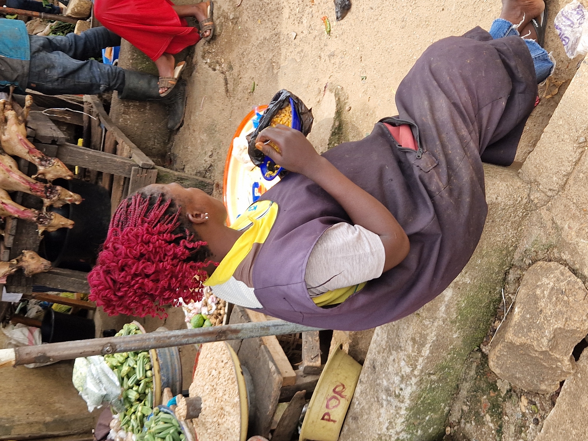 A person with bright red braided hair sits on a concrete ledge in an outdoor market in Kabong, Nigeria. They are wearing a dark purple smock over a grey t-shirt and blue jeans, and are eating from a black plastic bag held in a blue plastic bowl. The market appears busy, with piles of vegetables, grains, and meats displayed on wooden stands in the background. The ground is sandy and uneven, littered with rocks and debris. In the distance, other market-goers can be seen, along with a vendor with meat hanging from a wooden rack. The scene is brightly lit by natural daylight.