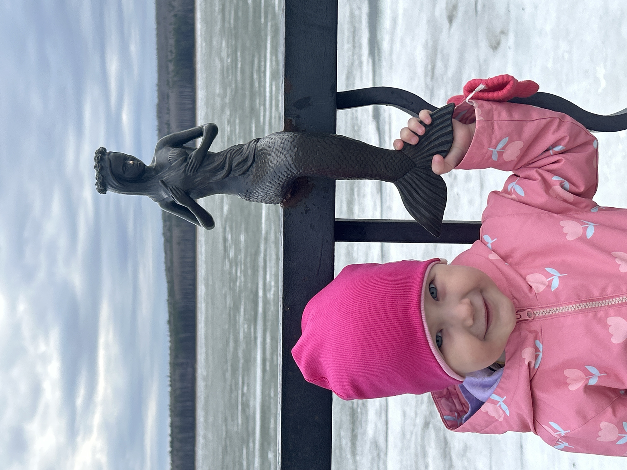 A close-up shot, rotated ninety degrees clockwise, features a young child in a pink hat and jacket holding the tail of a mermaid statue attached to a black metal railing. The child is smiling at the camera. In the background, a body of water stretches to the horizon, with trees visible along the far shore and a cloudy sky above. This scene appears to be from the city of Chaikovsky, Russia, as the mermaid statue is a known landmark in that city.