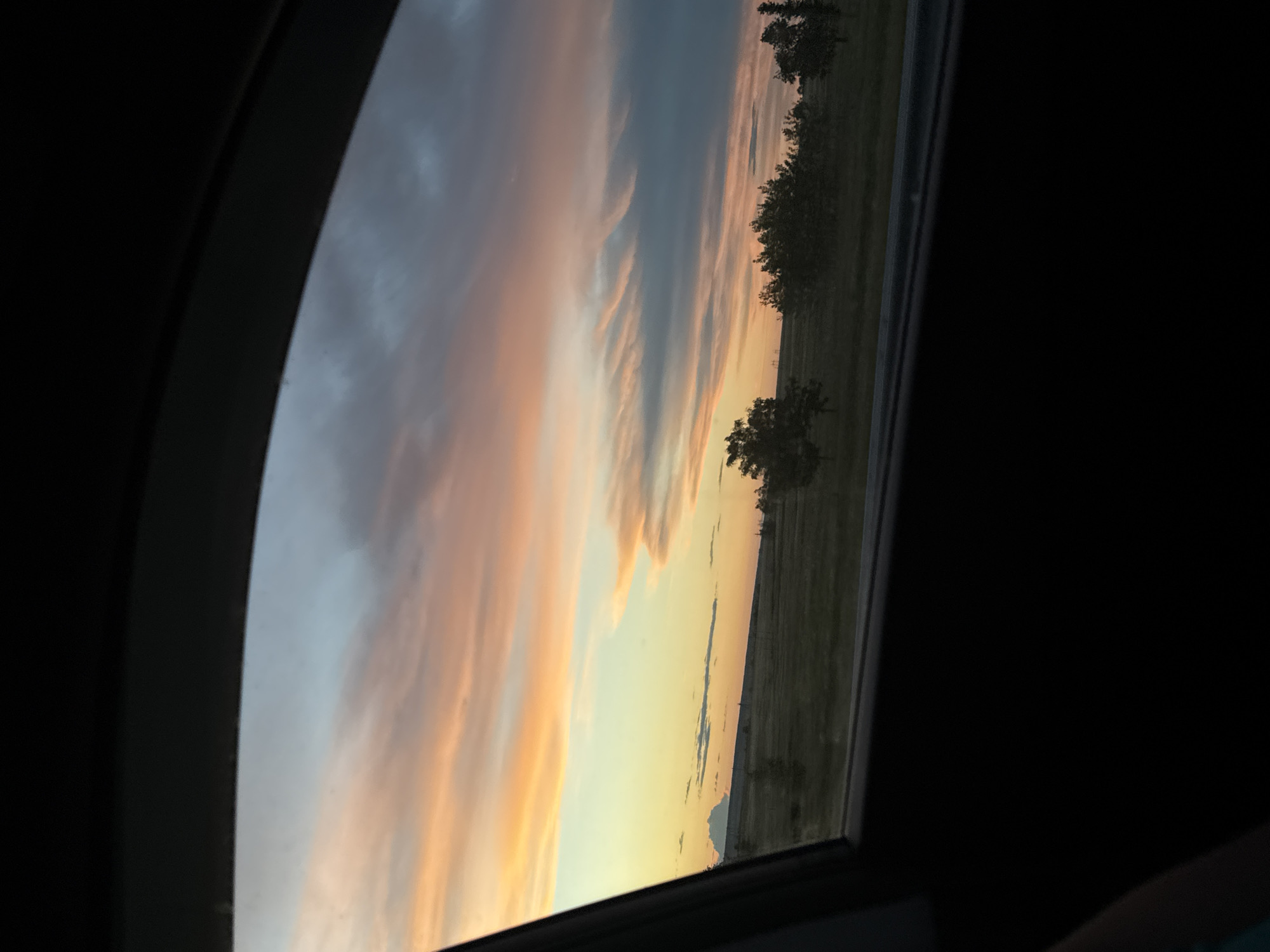 The image shows a view of a sky with clouds and a hint of sunset, seen through what appears to be a car window or sunroof. A thin strip of land with some trees is visible in the distance. The location is possibly in or near Ключи, Russia.