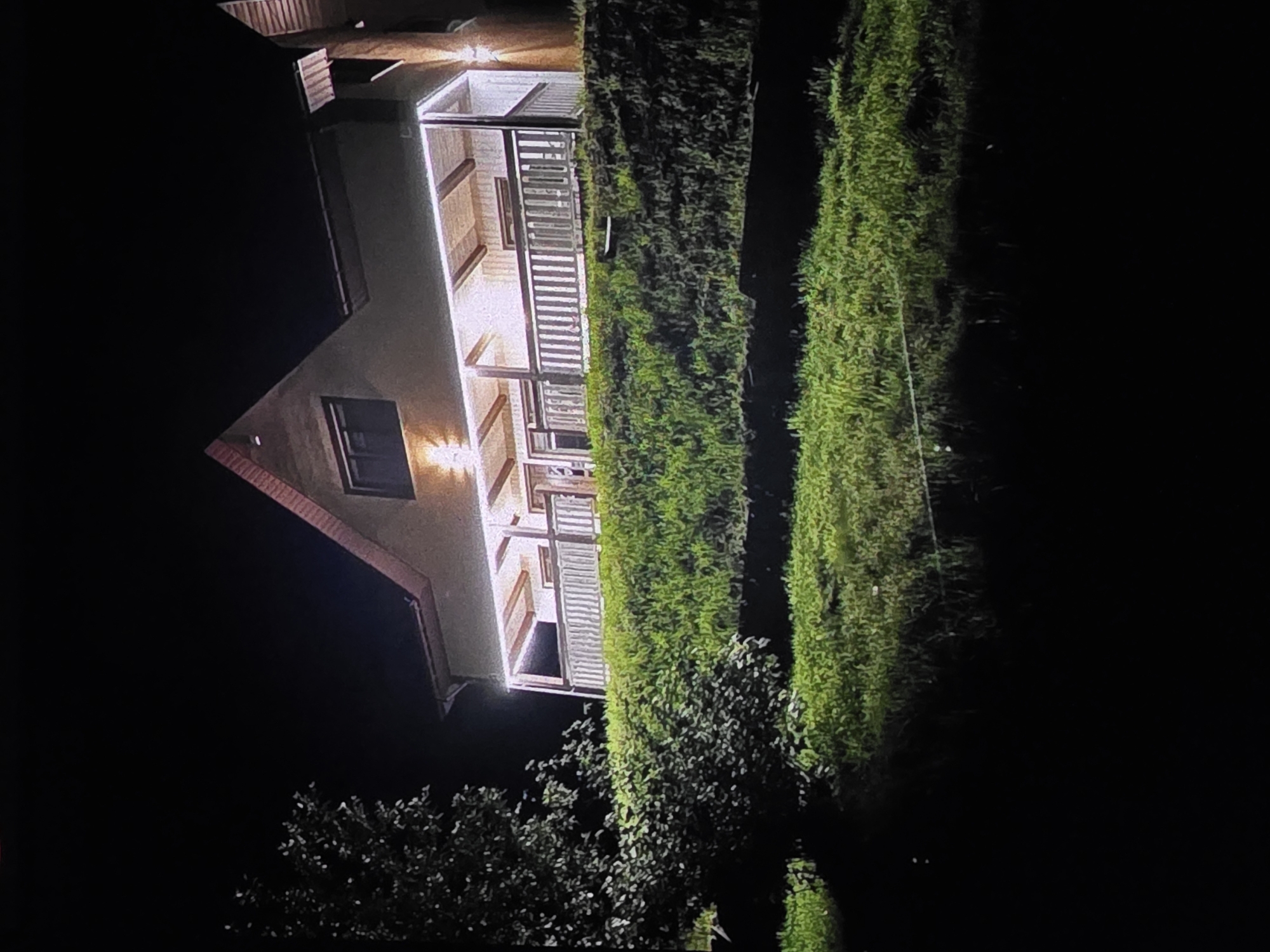 The image depicts a nighttime scene featuring a multi-story house with illuminated windows and a porch. The house has a light-colored facade and a red-tiled roof. Adjacent to the house are two tall, lush green hedges or trees, creating a strong vertical element in the composition. In the foreground, there are visible trees and foliage, which are darker and less distinct due to the low light conditions. The overall setting appears to be a residential area, possibly a garden or backyard, in Dubno, Ukraine. The time of day is clearly night, indicated by the darkness of the sky and the artificial lighting from the house. There are no people visible in the image, and the focus is on the architecture and the natural elements surrounding it. The lighting highlights the details of the house's porch, with visible wooden beams and railings. The tall hedges are well-lit by an unseen light source, creating a contrast with the dark surroundings. There is no visible text in the image. The scene conveys a sense of quietude and domesticity, with the illuminated house standing out against the night.