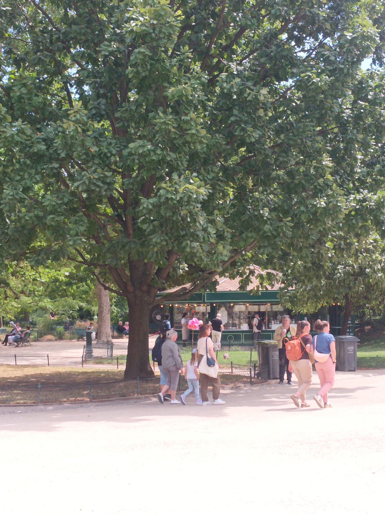 In Paris, France, a park scene unfolds under a large, leafy tree. In the foreground, three women walk across a light-colored pathway. The woman on the left wears a beige top, brown pants, and carries a beige tote bag. Next to her, a woman in a dark shirt and brown pants carries an orange backpack. The woman on the right is dressed in a blue denim shirt and pink pants, carrying a light-colored purse. A young girl in a white shirt and patterned pants walks ahead of them. In the midground, a kiosk with a green exterior and a brown roof is visible, with people browsing or working inside. Several trash cans are placed near the kiosk. Further in the background, a light-colored building or structure can be seen. People are seated on benches in the park, enjoying the day. The environment is characterized by lush green trees, grass, and pathways, typical of a Parisian park setting.