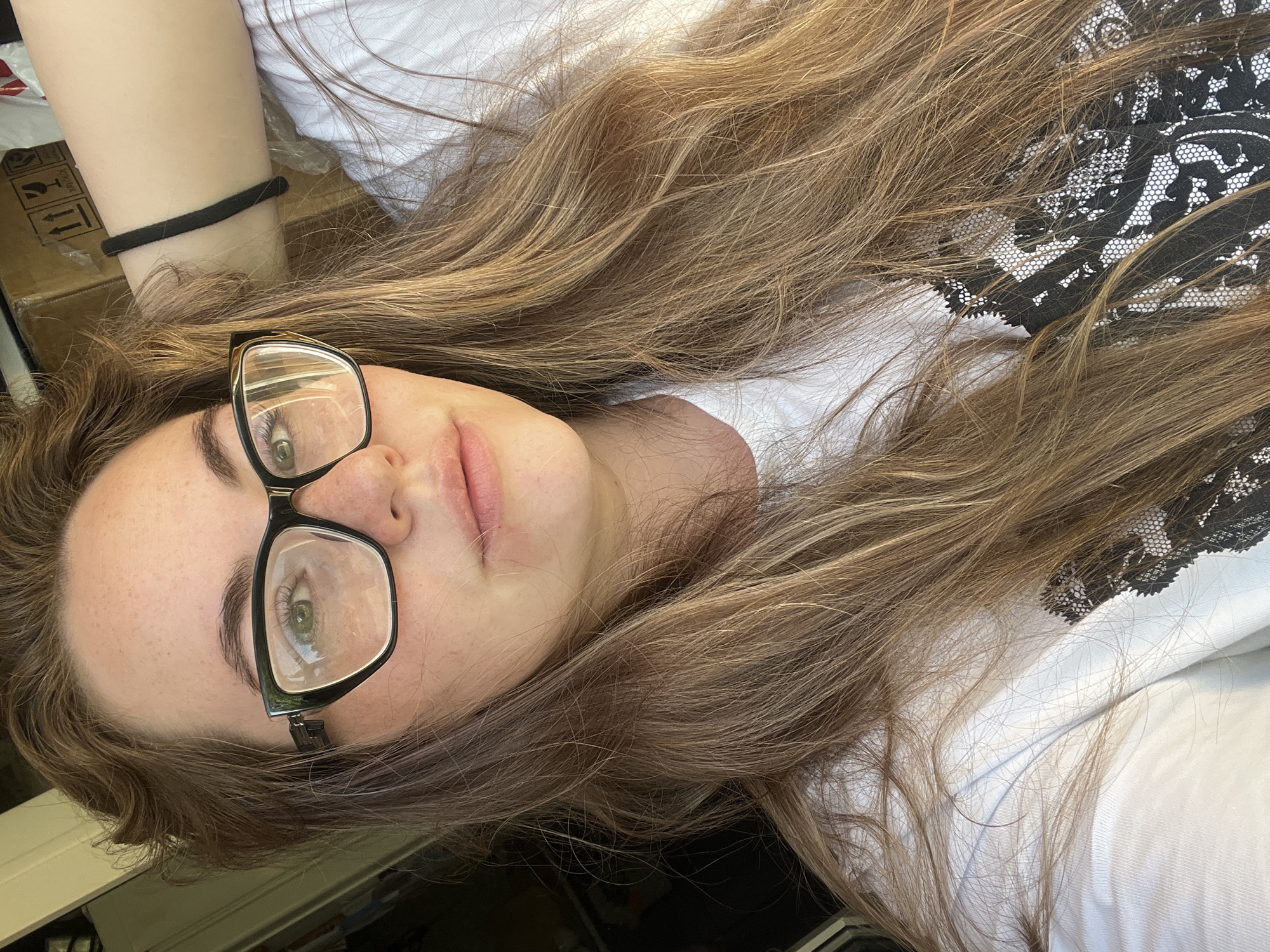 A woman with long brown hair and wearing glasses lies on her back. She is wearing a white t-shirt with black lace detailing. A black hair tie is visible on her arm. A cardboard box is in the background. The image lacks detail to ascertain it was taken in Chaikovsky, Russia.
