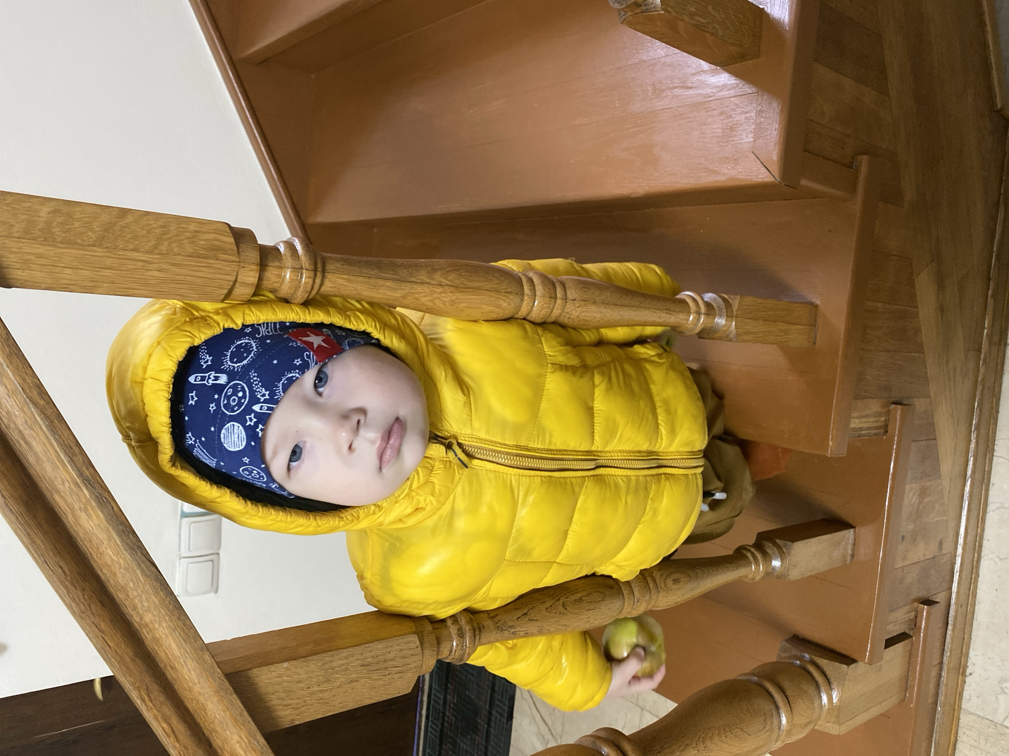 A single young child is depicted on a wooden staircase. The child wears a bright yellow hooded puffer jacket, zipped, over a dark blue beanie featuring white and red space-themed patterns, including a red star. The child has light-colored eyes and a neutral expression, looking directly at the camera. A green apple is held in the child's right hand. The child is positioned among the balusters of a wooden balustrade with a prominent handrail. The staircase has brown-stained wooden steps. The background consists of a plain light-colored wall with two white electrical outlets visible. The floor at the base of the stairs is light-toned. This image contains no discernible visual elements to confirm a location in the city of Sochi, Russia.