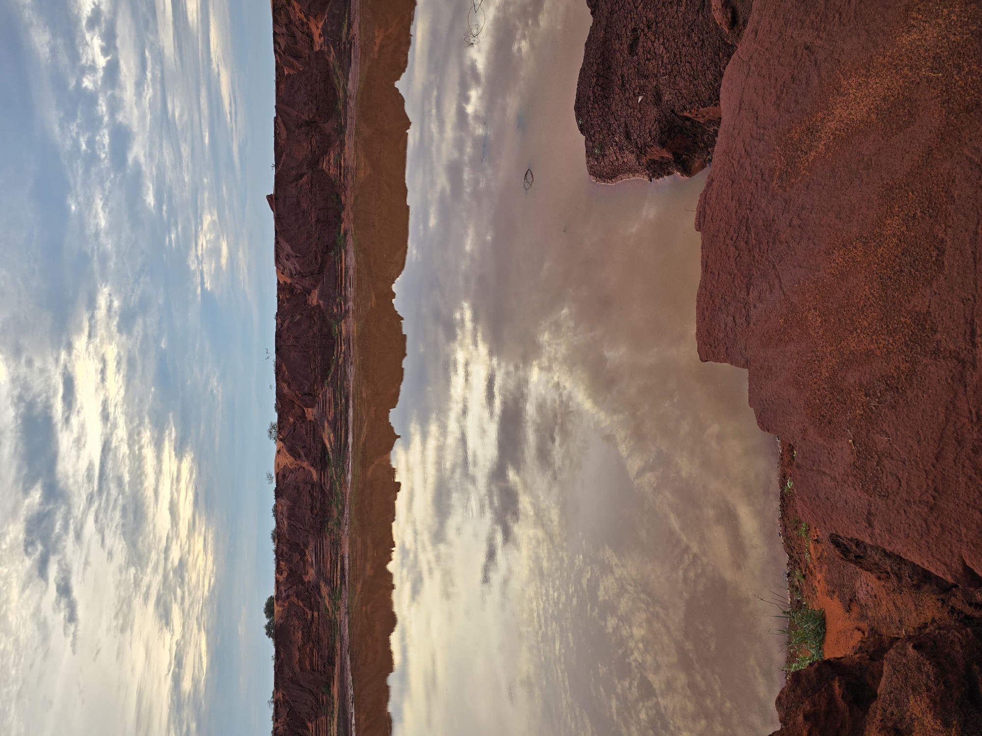 The image depicts a landscape in Mui Ne, Vietnam, featuring red rock formations, a body of water reflecting the sky, and cloud formations. The water's surface acts as a mirror, capturing the colors and patterns of the clouds. The reddish-brown rock formations are in the foreground and along the horizon and appear rugged. The sky shows varying hues of blue and white from the clouds. The photograph is taken from a low angle, emphasizing the height of the rock formations and the expansiveness of the sky. The scene lacks signs of human presence or activity.