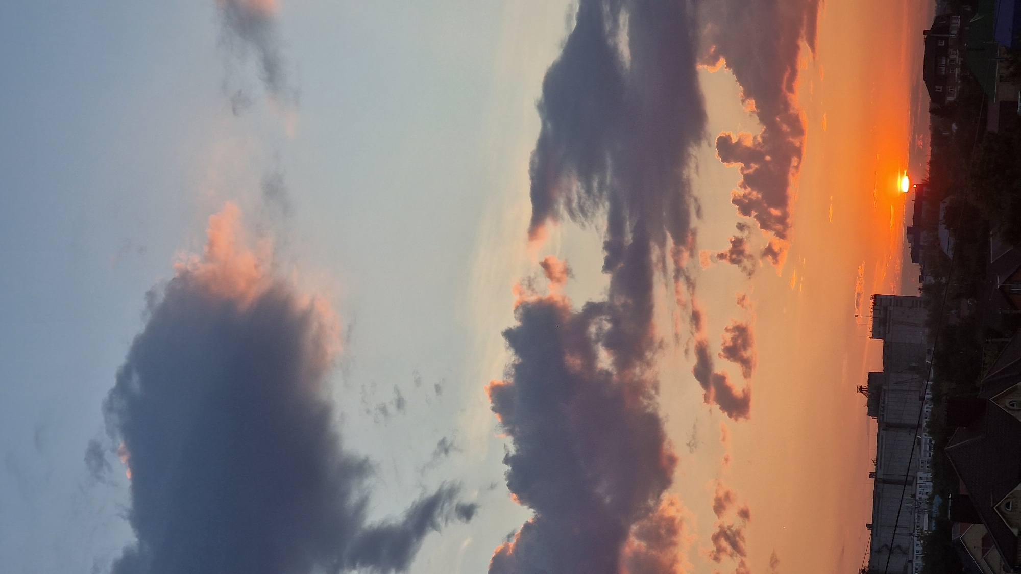 The image depicts a sunset over the city of Kostanay, Kazakhstan. A striking juxtaposition of elements is observed, with the warm hues of the setting sun illuminating the clouds and casting a glow over the urban landscape. In the foreground, residential rooftops are visible, suggesting a suburban or low-density urban area.
Dominating the middle ground is a large industrial-looking building complex, possibly a silo or factory, silhouetted against the vibrant sky. The architecture appears functional and utilitarian, with multiple structures of varying heights and designs. The sunset's rays highlight the textures and forms of this industrial zone.
The sky itself is a canvas of dramatic color. Dark, imposing clouds, tinged with pink and orange from the sunset, drift across the sky. These are interspersed with lighter, more wispy clouds, and areas of clear, pale blue sky towards the top of the frame. The sun, a brilliant orb, is partially obscured by the horizon and the clouds, indicating it is either just setting or has just set.
The overall impression is one of a typical urban sunset, where natural beauty intersects with man-made structures. No people or animals are discernible in the image, and there are no overt actions or interactions taking place. The focus is entirely on the atmospheric conditions and the urban environment bathed in the twilight glow. The scene suggests a moment of transition, from day to night, painted with the dramatic colors of a sunset.
