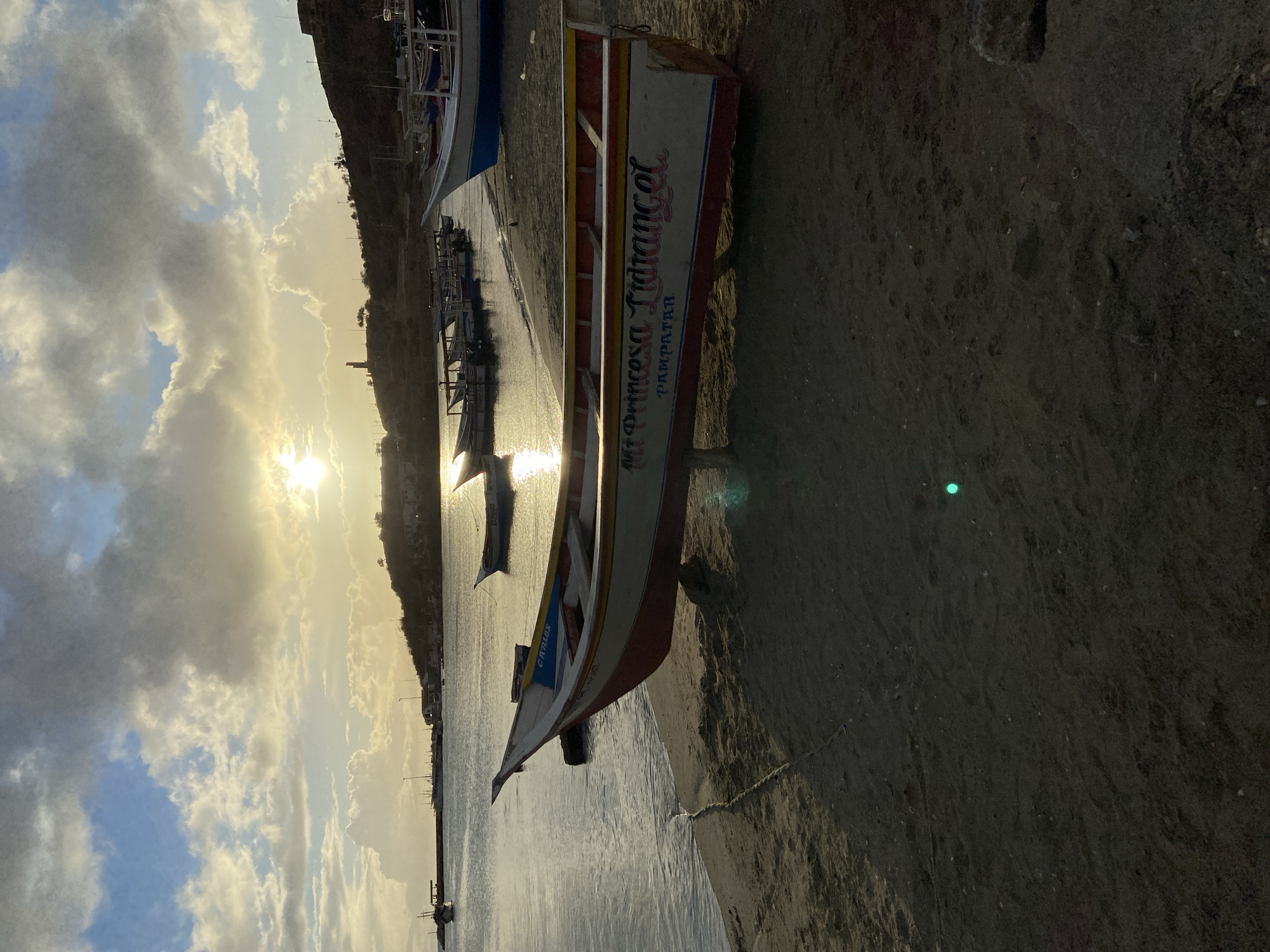 The image depicts a sunset scene at the bay of Juan Griego, Venezuela. A wooden boat named "Mi Princesa Lidiangel" is beached on the sand in the foreground, its hull painted white with red and blue accents. The boat's interior shows wooden benches and bracing. In the background, several other fishing boats are moored in the calm water. The sun, low on the horizon, casts a warm glow over the scene, reflecting on the water and illuminating the clouds. The shoreline is a mix of sand and some rocky debris. A hillside with scattered buildings and a prominent tower can be seen in the distance, silhouetted against the sky. The overall atmosphere is peaceful and serene, characteristic of a coastal town at dusk.