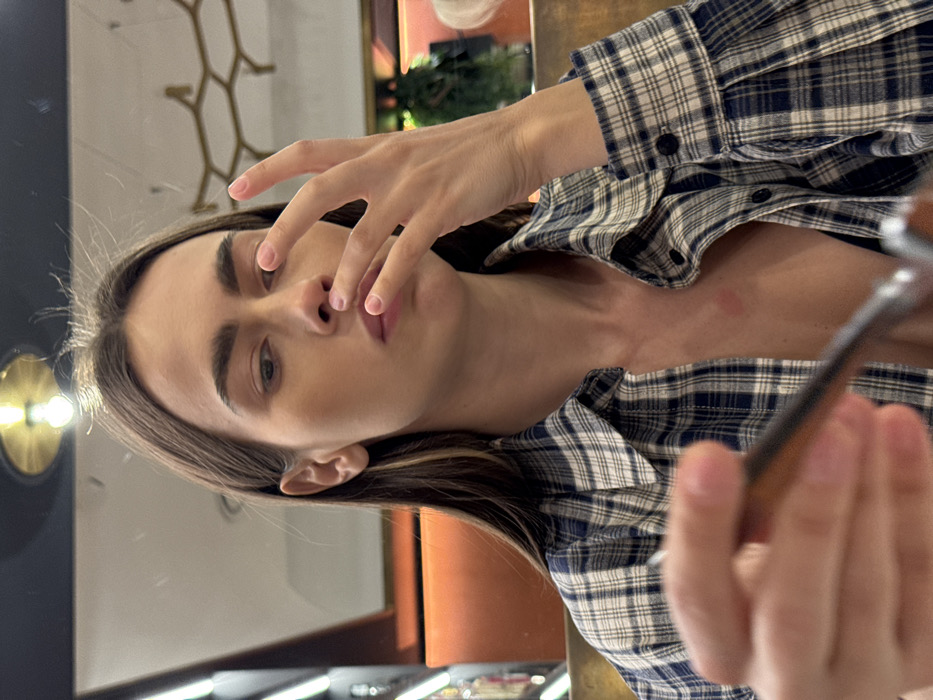 An indoor scene in Verona, Italy, depicts a young woman from the chest up, positioned slightly off-center. She has long, dark brown hair with lighter strands, fair skin, and prominent eyebrows. Her left hand, with light pink nail polish, is raised with fingers touching the area between her nose and mouth. Her eyes are cast downwards or to her right. She wears a blue and white plaid button-up shirt, partially unbuttoned, revealing a dark undergarment. A distinct reddish mark is visible on her upper chest, near her left collarbone. In the blurred foreground, her right hand holds an out-of-focus object, possibly a spoon handle or eyewear. The background suggests a modern commercial establishment, likely a cafe or bar. Visible elements include a circular overhead light fixture with a gold rim reflected in a dark, possibly mirrored ceiling. To the right, a gold-colored, geometric, honeycomb-like wall or ceiling decoration is present. On the left, vertical strip lighting and an orange-brown wall panel are partially visible. The overall lighting is warm.