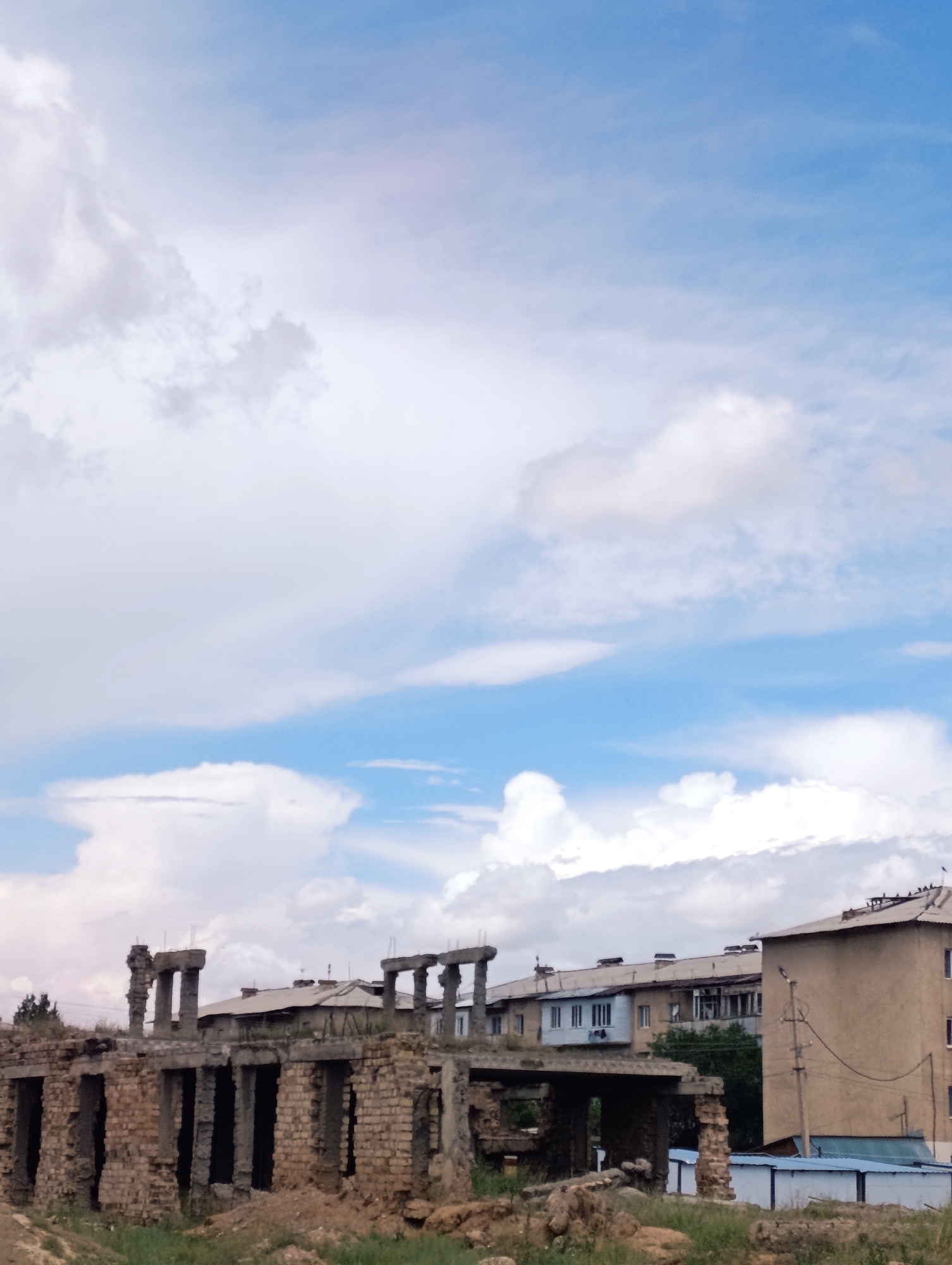 In Naryn City, Kyrgyzstan, an outdoor scene captures the contrast between a partially constructed or ruined building in the foreground and a residential building and apartment complex in the background. The foreground features a weathered structure made of brick and concrete, with exposed columns and incomplete walls. The ground surrounding this structure is rough and covered with dirt and sparse vegetation.
Behind the ruined building, a light-colored apartment building stands, its facade marked by windows and balconies. Further in the background, a multi-story residential complex with a sloped roof is visible, with multiple windows and rooftop chimneys. The sky above is a mix of blue and white, with scattered clouds. There are no people visible in the image. The overall impression is one of an area undergoing construction or experiencing decay, juxtaposed with ongoing urban life.