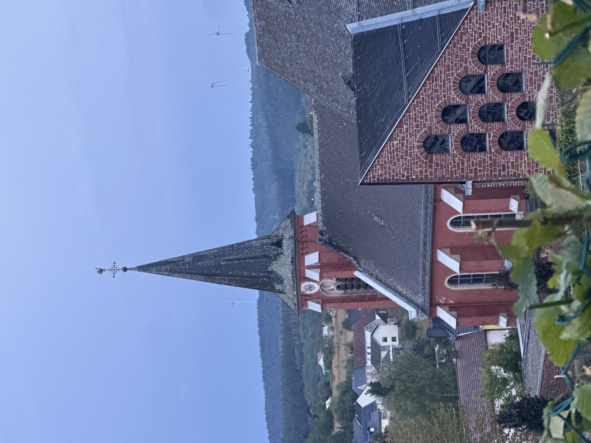 The image depicts the parish church of St. Martin in Wadern, Germany. The church features a tall, dark steeple topped with a weather vane shaped like a rooster. The main body of the church is painted red and has arched windows. Adjacent to the steeple is a section of the church constructed from red brick, with a series of smaller arched windows. In the background, a village with houses and trees is visible, situated on a hillside. Further in the distance, a dense forest covers rolling hills, and several wind turbines can be seen on the horizon. The sky is overcast with a pale blue hue. There are no people or animals visible in the image.