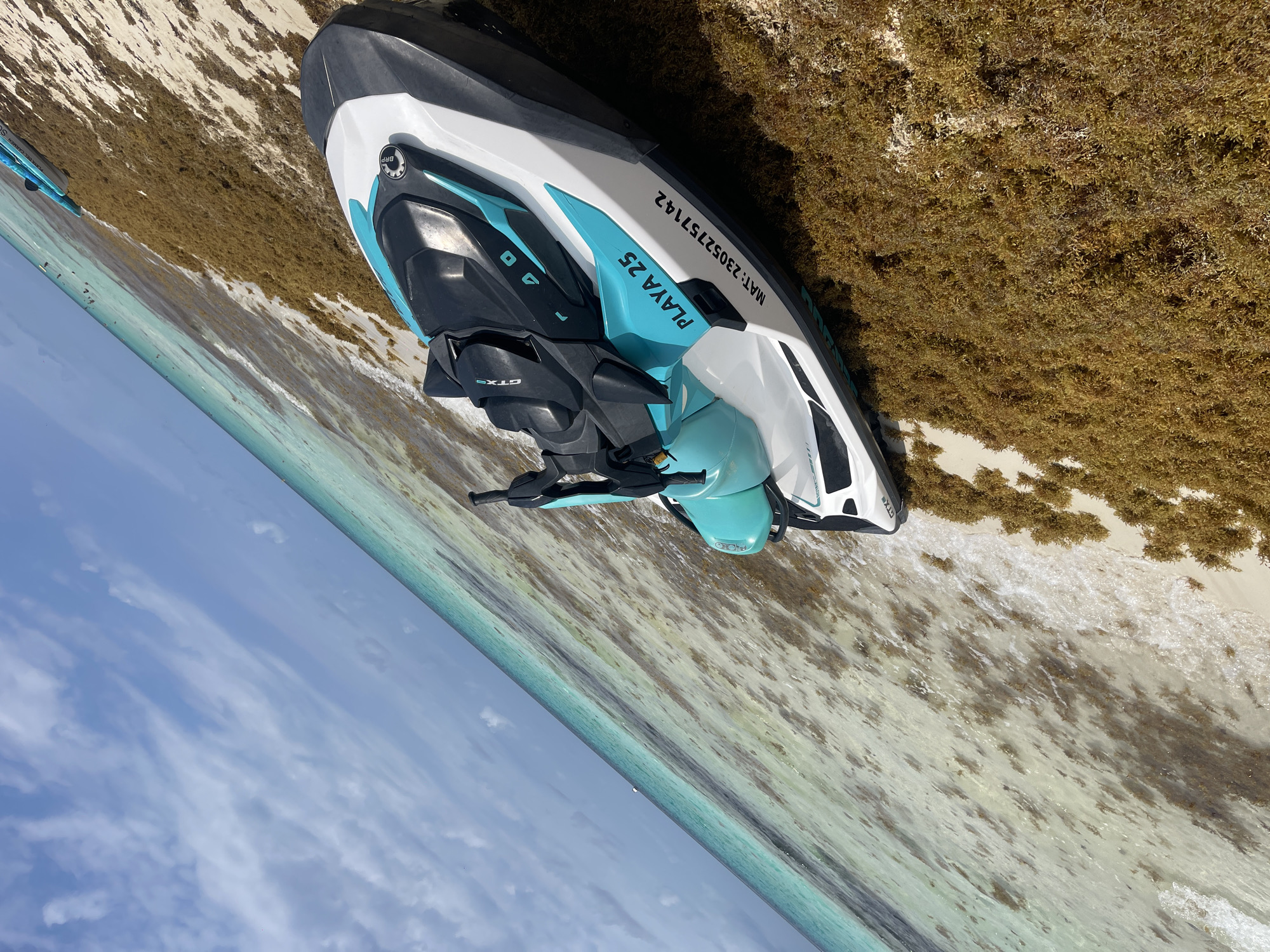 A jet ski is parked on a beach next to the ocean. The jet ski is white, blue, and black, with "PLAYA 25" printed on its side. The license plate number MAT: 23052757142 is also on the side of the jet ski. The beach appears to be covered with seaweed. The ocean is a clear turquoise color. The sky is blue with some clouds. The scene appears to be in Playa del Carmen, Mexico, based on the text on the jet ski.